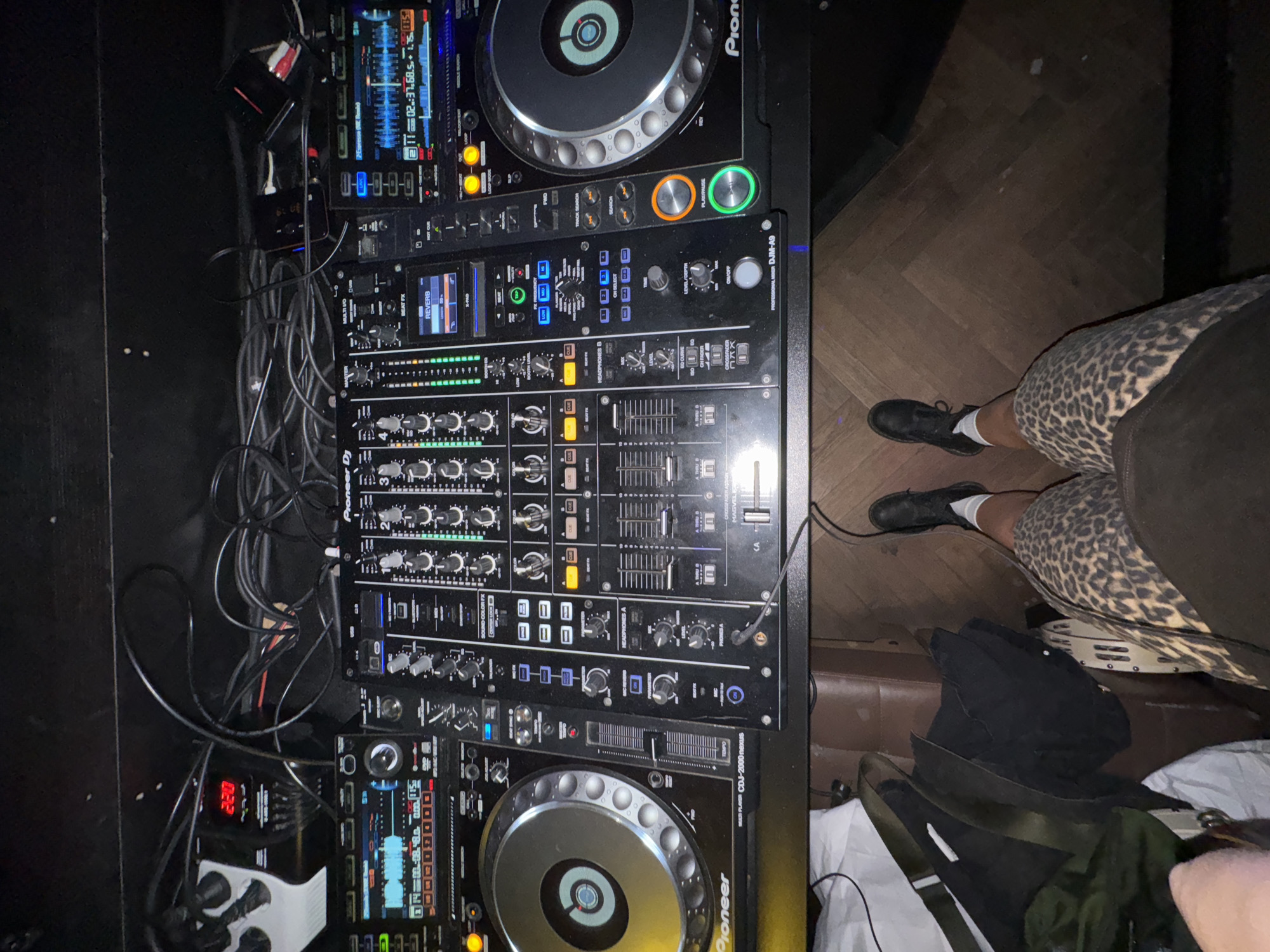 The scene depicts a DJ setup in Moscow, Russia, viewed from a first-person perspective looking down. The central focus is a professional DJ console comprising two Pioneer CDJ-2000nexus multi-players flanking a Pioneer DJM-A9 professional mixer. The left CDJ-2000nexus displays waveform data and track information including "11 00.58.040" and a tempo of "115" BPM. The right CDJ-2000nexus also shows waveform data and track information, specifically "11 02.37.68.S." with a pitch adjustment of "+1.5." The DJM-A9 mixer features four channels, various sound color and beat effects, with "REVERB" currently selected. A headphone cable is plugged into the mixer's "HEADPHONES A" jack.
Above the equipment, an array of black cables is visible, connected to a white power strip on the far left. A small black digital display shows "220," indicative of the voltage. The floor beneath the console is constructed of dark brown wooden planks arranged in a herringbone pattern. A person is standing in front of the equipment, their legs visible wearing dark lace-up boots, white socks, and leopard-print shorts or a skirt. A dark bag with a green strap and a white cloth are positioned to the left of the person's feet.