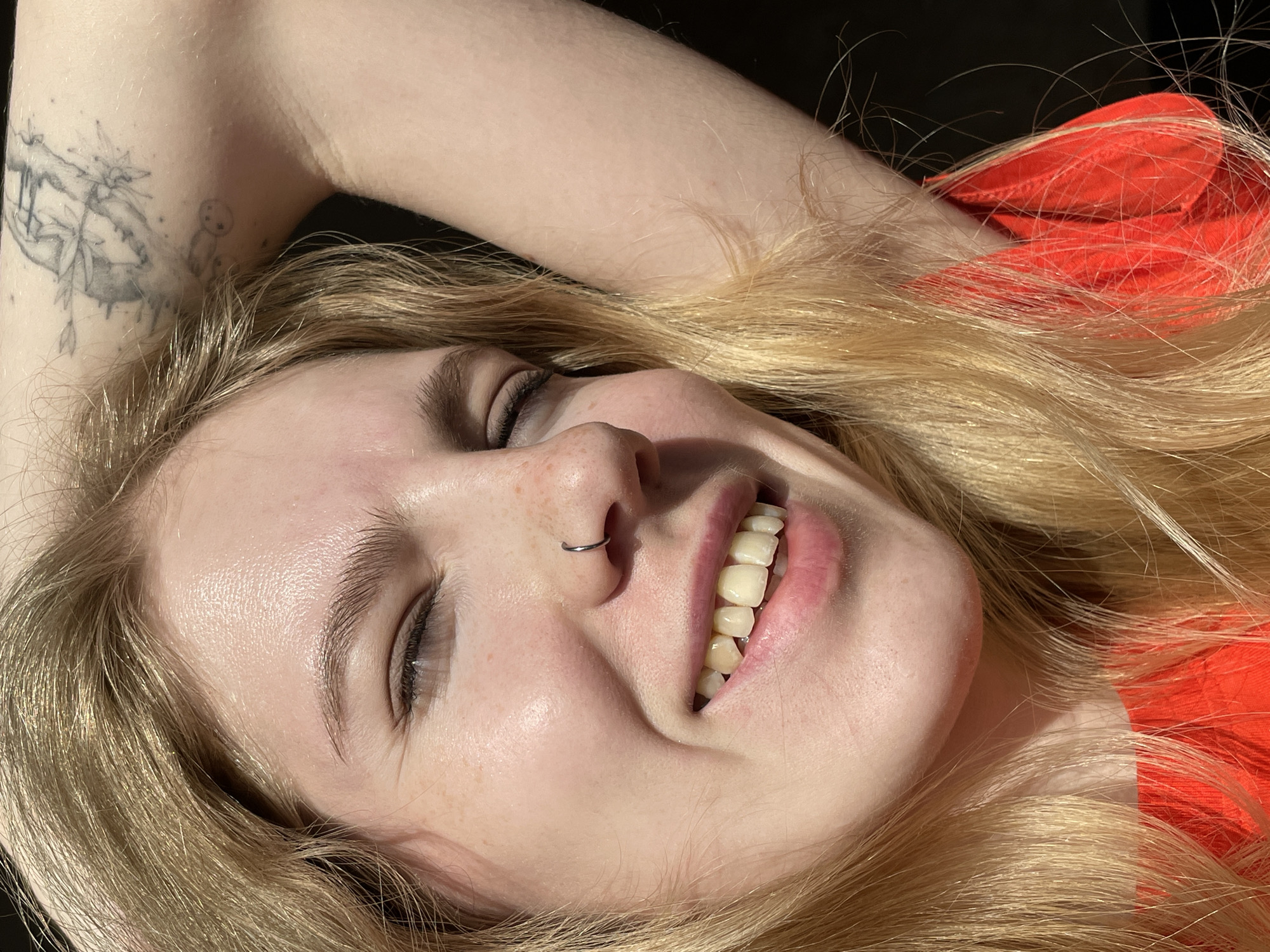 The image shows a close-up, selfie-style shot of a young blonde woman with her right arm raised and hand behind her head, her eyes are closed and she is smiling. She has a silver hoop nose ring and several small freckles on her cheeks. She is wearing an orange top. A black tattoo is visible on her upper arm. The lighting suggests a bright, sunny day. The background is dark and out of focus. There is no indication that this image was taken in Izhevsk, Russia or any specific city.