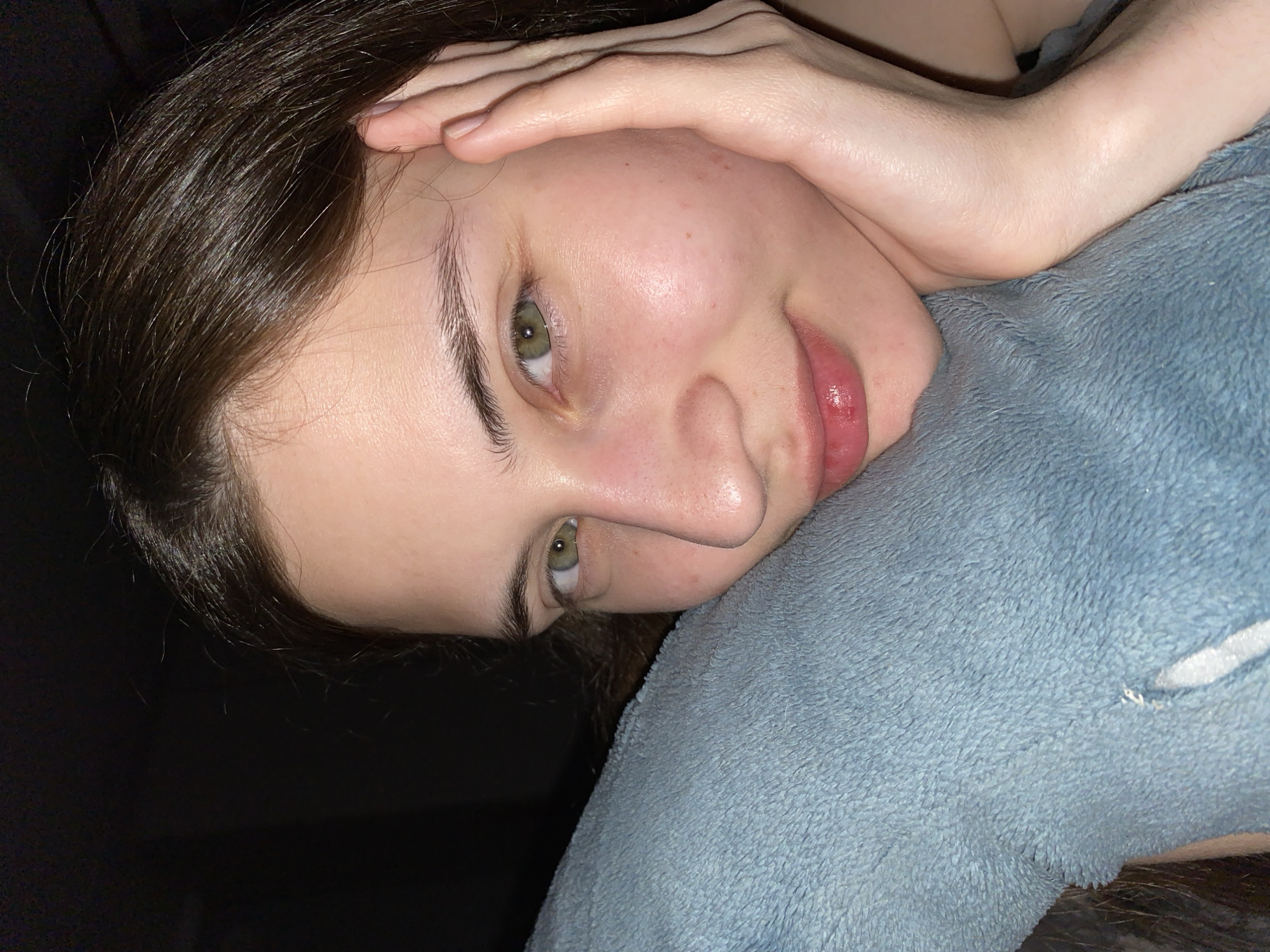 A young woman with dark hair and green eyes is lying on a soft, blue pillow. Her hand rests on her forehead. The background is dark and indistinct. There is no evidence suggesting the location is in Votkinsk, Russia.