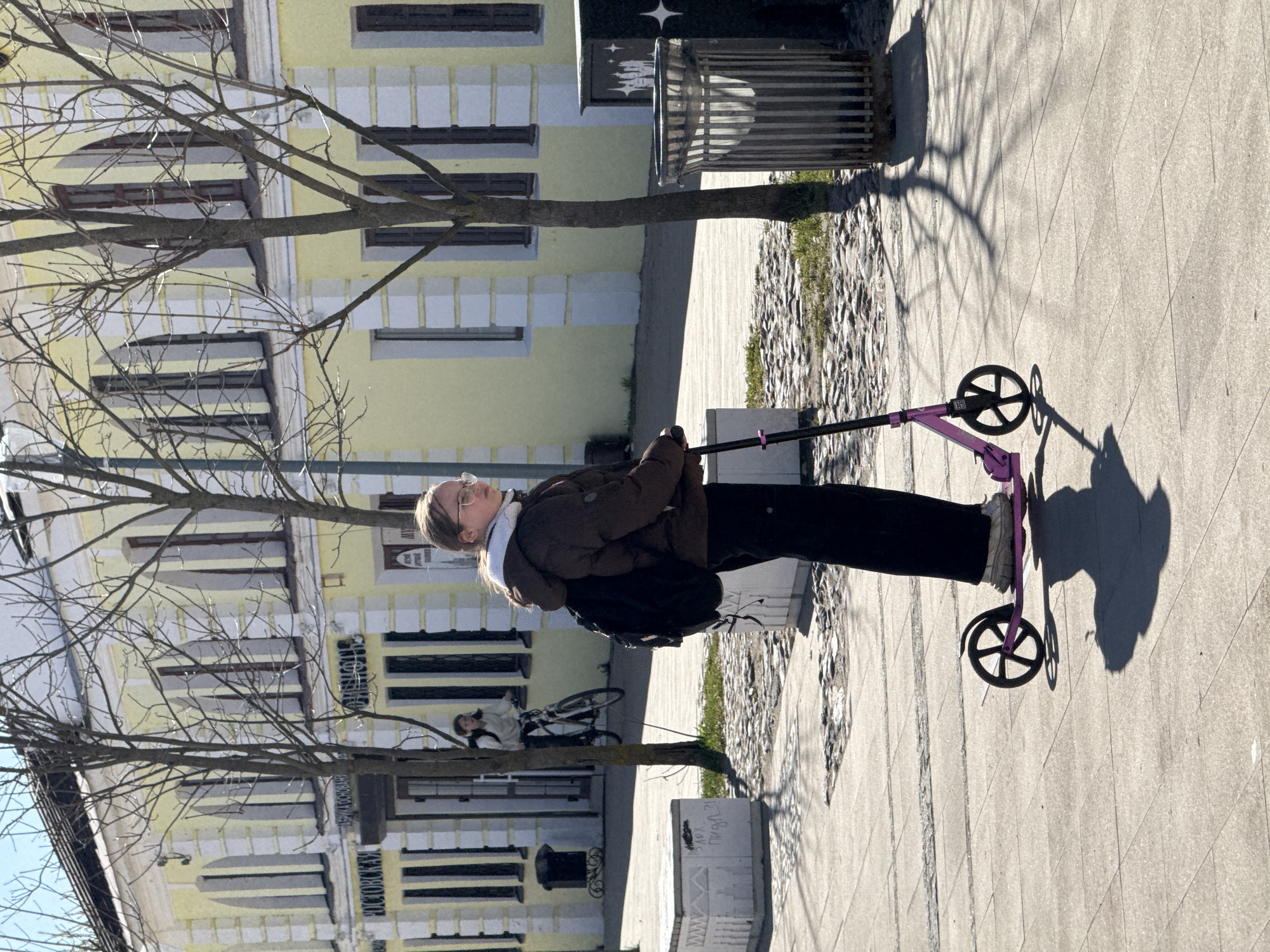 A person rides a purple scooter on a sidewalk in Ново-Никольское, Russia, alongside a yellow building with many windows. The person wears a brown jacket, black pants, and sneakers. They also have a white scarf and glasses. A black backpack is slung over their shoulder. A bare tree is in the foreground, casting shadows on the sidewalk. In the background, a person leans against a bicycle near the building. A metal trash bin and a concrete block are also visible.