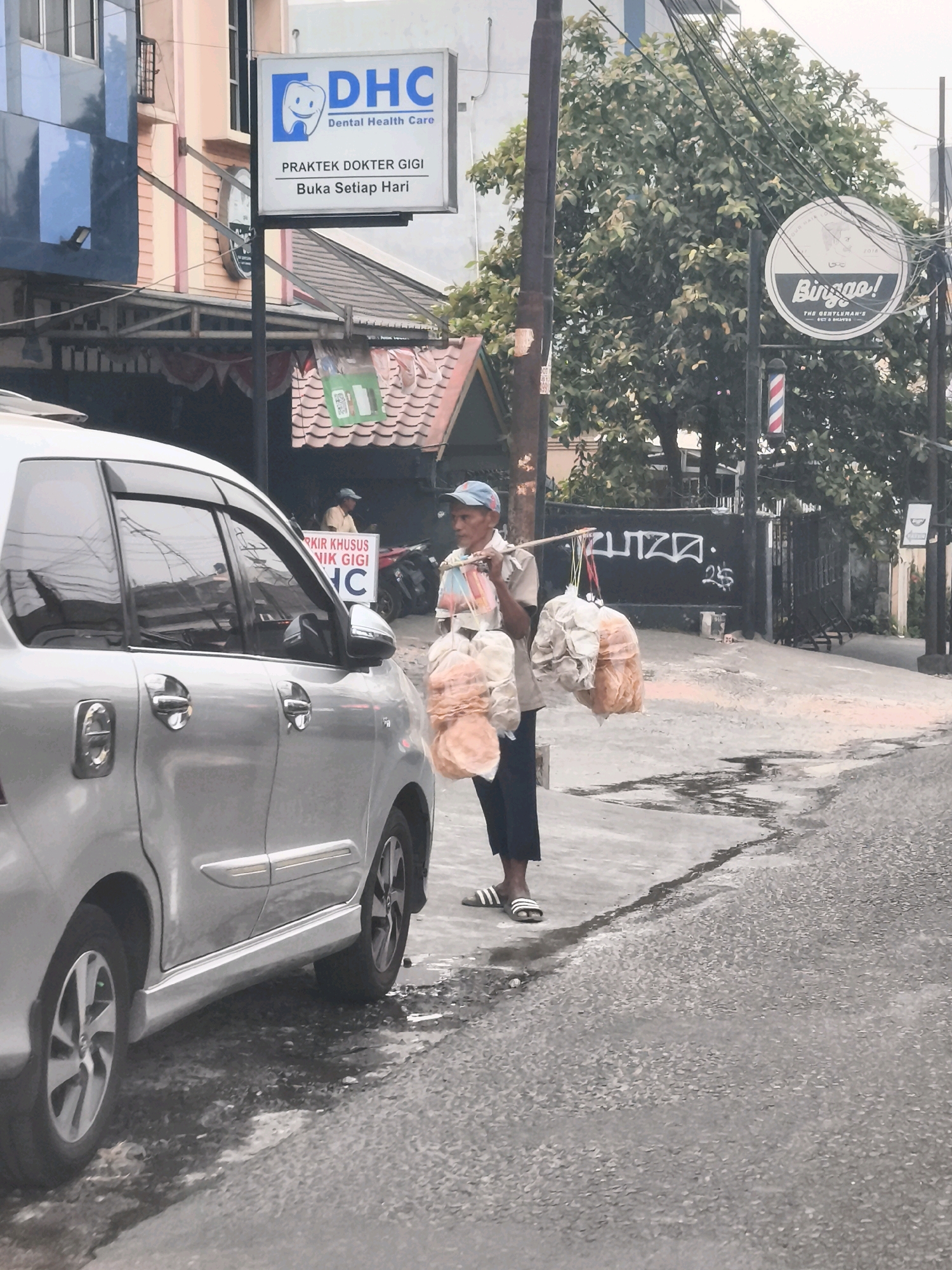 In Palembang, Indonesia, a street vendor carrying bags of snacks on a shoulder pole walks past a silver minivan on a paved road. The vendor, wearing a blue cap and a light-colored shirt with dark pants, is walking from left to right in the frame. He is carrying three bags of snacks, two of which are large and transparent, filled with what appear to be puffed snacks, and a smaller bag in front. The minivan is parked on the left side of the road, facing away from the viewer. Behind the vendor and minivan, a building with signs is visible. One sign, in English and Indonesian, reads "DHC Dental Health Care" with the practice hours indicated below. Another circular sign, "Binggo! THE GENTLEMAN'S BARBER SHOP," is also visible. A red and white striped barber pole stands next to it. The road appears wet in places, suggesting recent rain. Trees and utility poles are also present in the background.