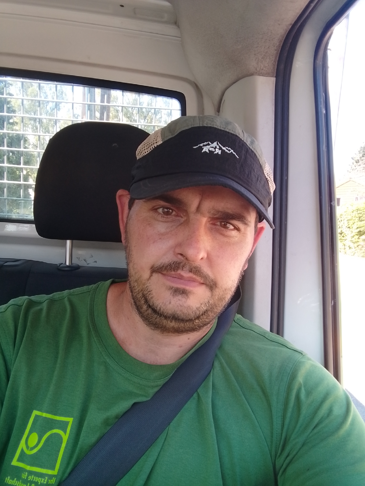 A man wearing a green t-shirt and a black and grey baseball cap with a mountain logo is sitting in a vehicle. He has short brown hair and a beard, and he is looking at the camera. The man is wearing a seatbelt. The interior of the vehicle is visible behind him, with a headrest and part of a window. Outside the window, trees and a building can be seen. The image appears to be a selfie taken inside the vehicle. The image is not geotagged, so its location in Schenefeld, Germany, cannot be confirmed.