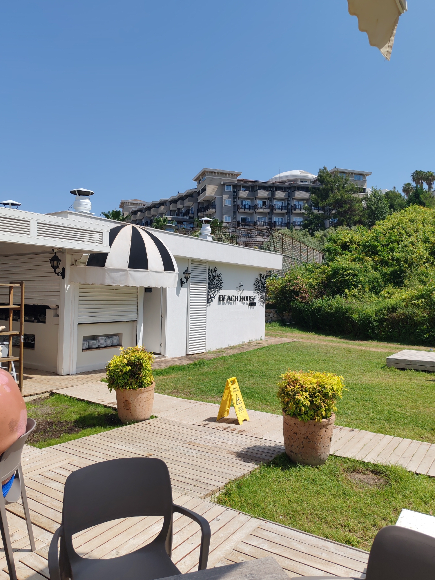 The image shows an outdoor area in Alanya, Turkey, likely a beachside establishment. A low, white building with a black-and-white striped awning stands to the left. The building's exterior is adorned with the words "BEACH HOUSE" and tree-like decorations. A closed shutter covers one side of the building. A yellow warning sign stands on a wooden walkway that extends from the building. Two large pots containing yellow plants flank the walkway. Modern black chairs are placed on the wooden deck, and a glimpse of a person's arm is visible in the foreground. In the background, a large, multi-story building with balconies and a distinctive dome-shaped roof is situated on a hill partially obscured by lush greenery. The sky above is a clear, bright blue.