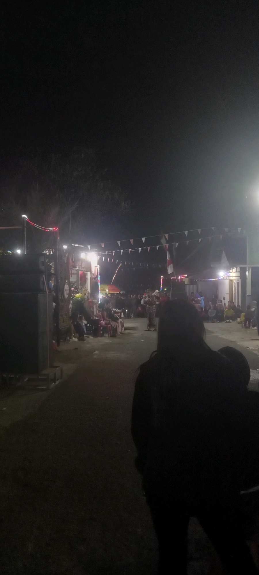 The image depicts a nighttime outdoor gathering in a village setting, likely in Argasoka, Indonesia. The scene is illuminated by various lights, including string lights with red and white bulbs, and brighter lights emanating from what appears to be a building or stall in the background. A line of triangular flags, some red and white, stretches across the scene, suggesting a celebratory or festive event.
In the foreground, a person is silhouetted against the lights, obscuring details of their appearance but suggesting their presence as an observer or participant. Further into the scene, there are groups of people seated on chairs and standing. Some individuals are visible in the mid-ground, and a larger crowd is gathered in the background, attending to an activity or performance that is not clearly discernible.
To the left of the image, large dark objects, possibly speakers or sound equipment, are positioned. The ground appears to be a dark, paved or dirt path. The overall atmosphere is that of a community event held in a local, possibly residential or communal area, at night. The lighting creates a somewhat hazy effect, characteristic of outdoor night photography.