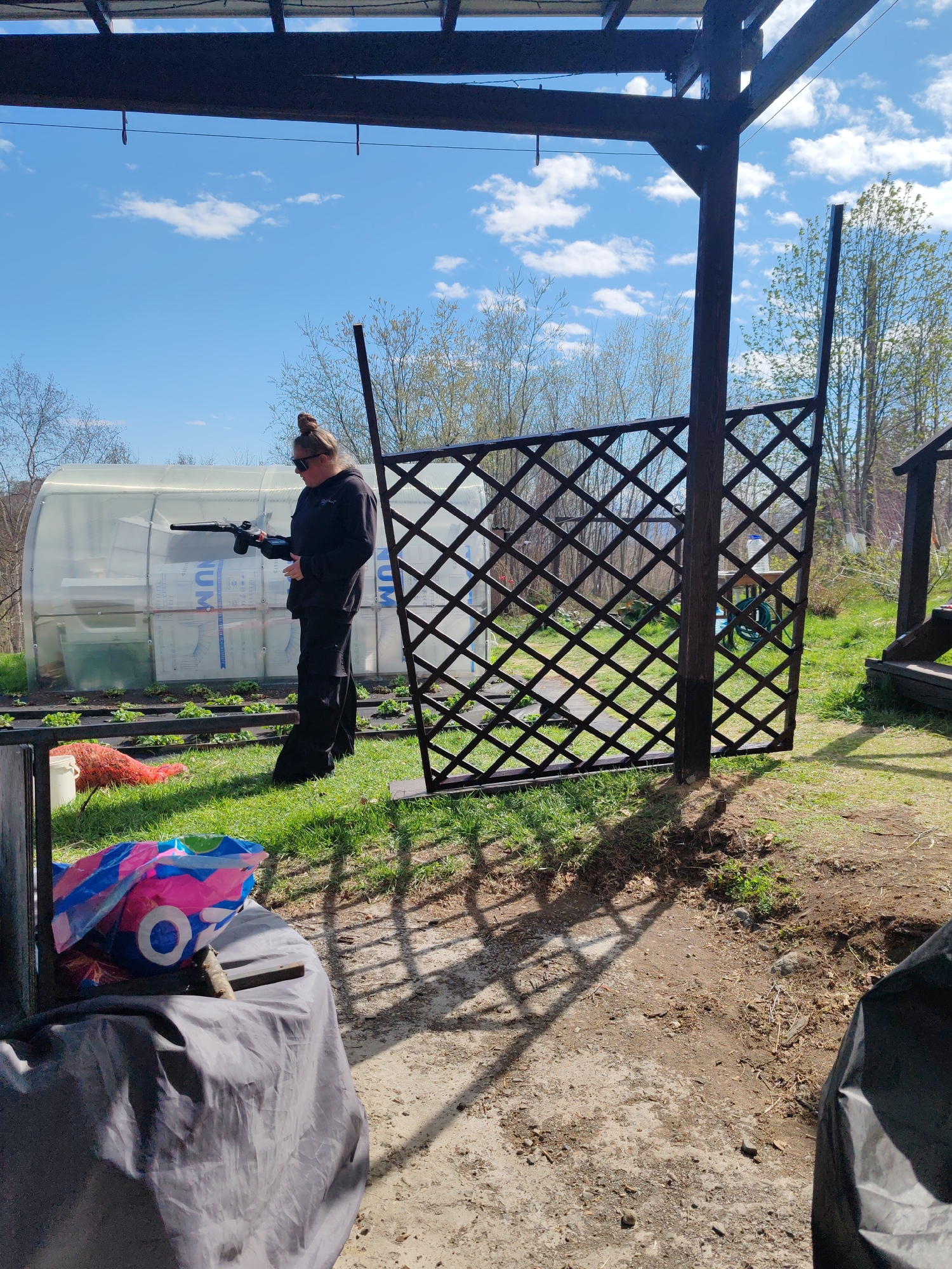 A woman in black clothing and sunglasses stands on a grassy lawn, holding a dark-colored object resembling a pump-action pistol or air rifle. Behind her is a semi-transparent greenhouse-like structure, and a dark brown, open lattice fence panel supported by a dark wooden post stands to her right. A covered object and a colorful bag are visible in the foreground on a dirt patch. The setting appears to be in a backyard or garden area, with trees and a blue sky with scattered clouds visible in the background. The image does not contain sufficient information to determine that it was taken in Koryaki, Russia.