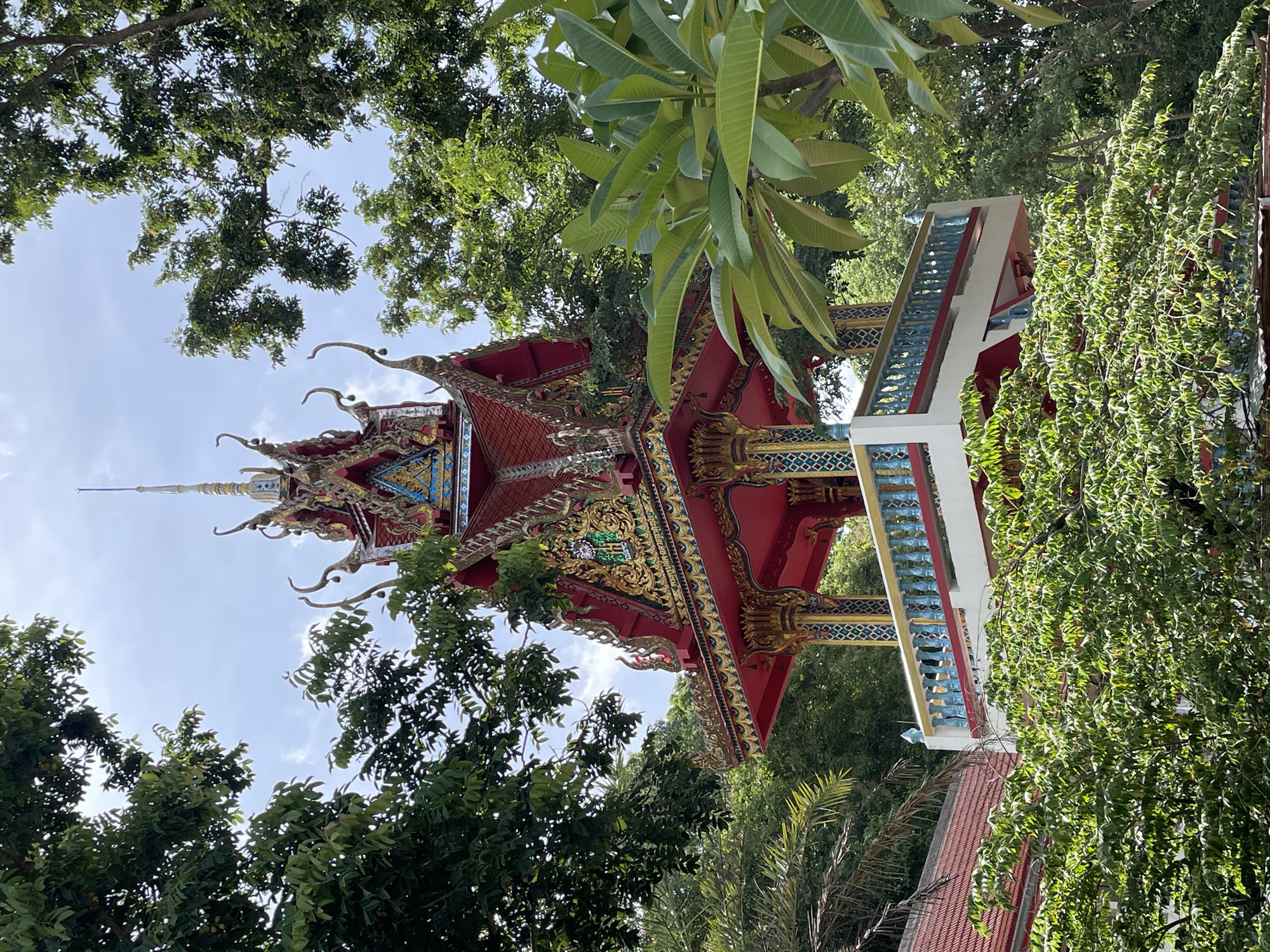 This is a vertical, eye-level, full shot of a multi-tiered, ornate temple tower surrounded by lush green foliage. The tower features a red, multi-gabled roof adorned with intricate gold and blue decorative elements and ornate finials. The middle section of the tower is open, with a white platform and a decorative blue and gold balustrade. The structure is supported by red and gold pillars. Green trees and palm fronds frame the tower, with a clear, lightly clouded sky visible above. The image appears to have been taken in Pattaya City, Thailand, given the architectural style common to Thai temples.