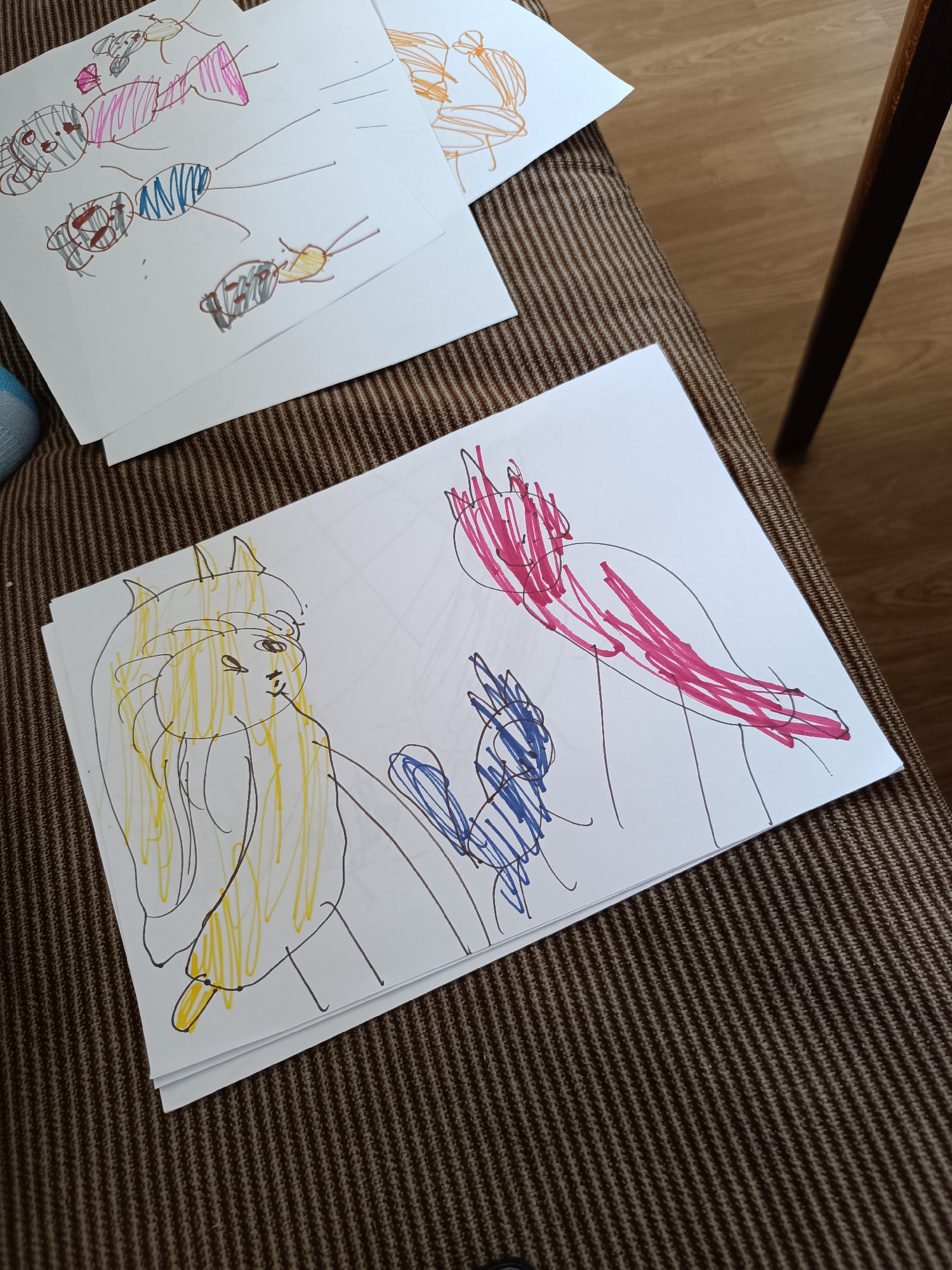 The image shows several sheets of paper with children's drawings on a brown, textured surface, possibly a couch or chair. In the foreground, a stack of papers features a drawing of three figures: one with yellow hair and a face, one with blue scribbles, and another with red scribbles. In the background, two additional drawings are visible, one with pink and brown figures, and another with orange scribbles. The location is unspecified.