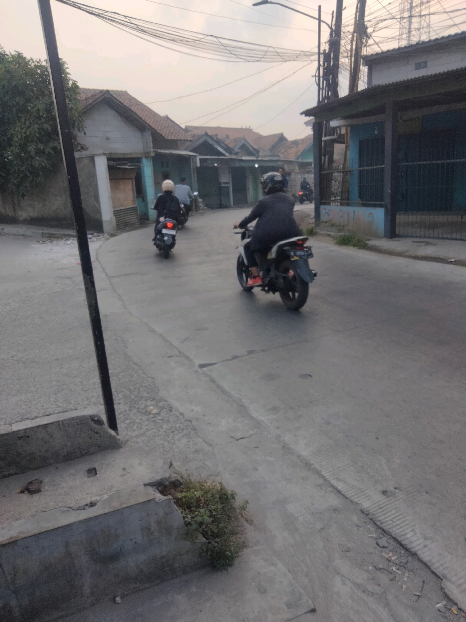 In Pisangan Jaya, Indonesia, a scene unfolds on a paved street lined with modest houses and small shops under an overcast sky. A dark-haired individual wearing a black jacket and helmet rides a white motorcycle, its rear brake light illuminated, down the street. Another motorcycle, ridden by someone with a beige helmet, travels ahead and to the left. The road shows signs of wear with cracks and a distinct demarcation in the pavement. A dark pole stands prominently on the left side of the image, and to the right, buildings with corrugated metal roofs and blue-painted facades are visible. Some vegetation sprouts from the side of the street. The overall impression is one of a typical day in a residential urban environment.