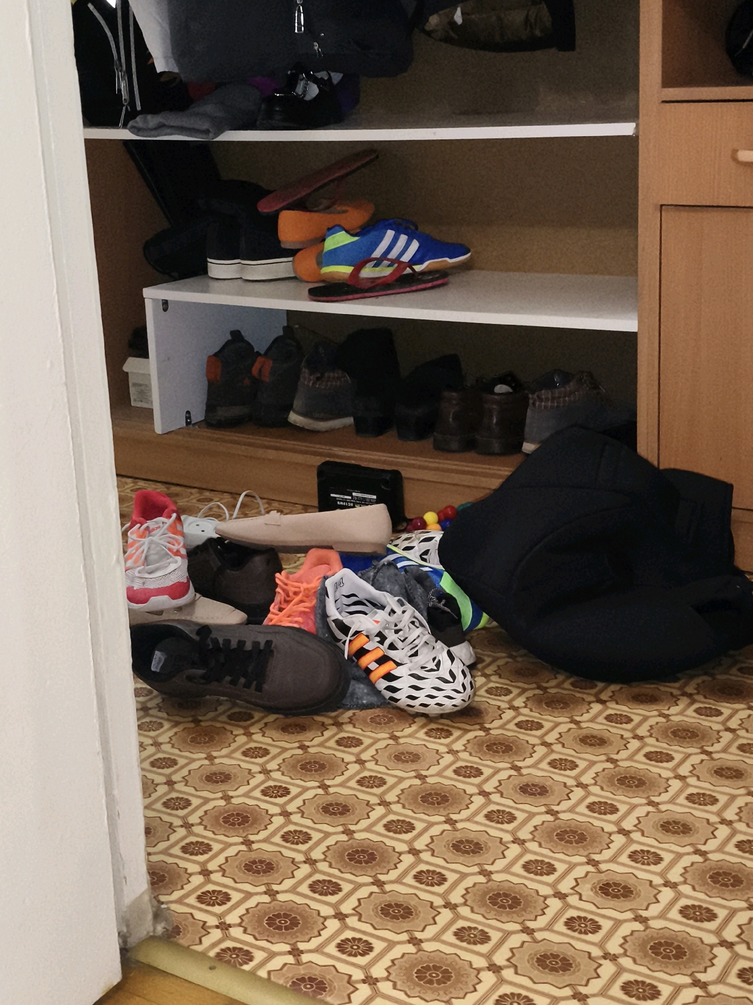 The image shows a closet filled with shoes and bags. The closet has several shelves. On the top shelf, there are black bags and some clothing. On the second shelf, there are black and white shoes, blue and yellow sneakers, and orange and black flip-flops. On the bottom shelf, there are black and gray shoes. On the floor in front of the closet, there are more shoes, including pink and white sneakers, beige flats, orange sneakers, and black and white sneakers with orange stripes. A large black bag is also on the floor. The floor is covered with a patterned tile. The scene is indoors, likely in a residential setting.