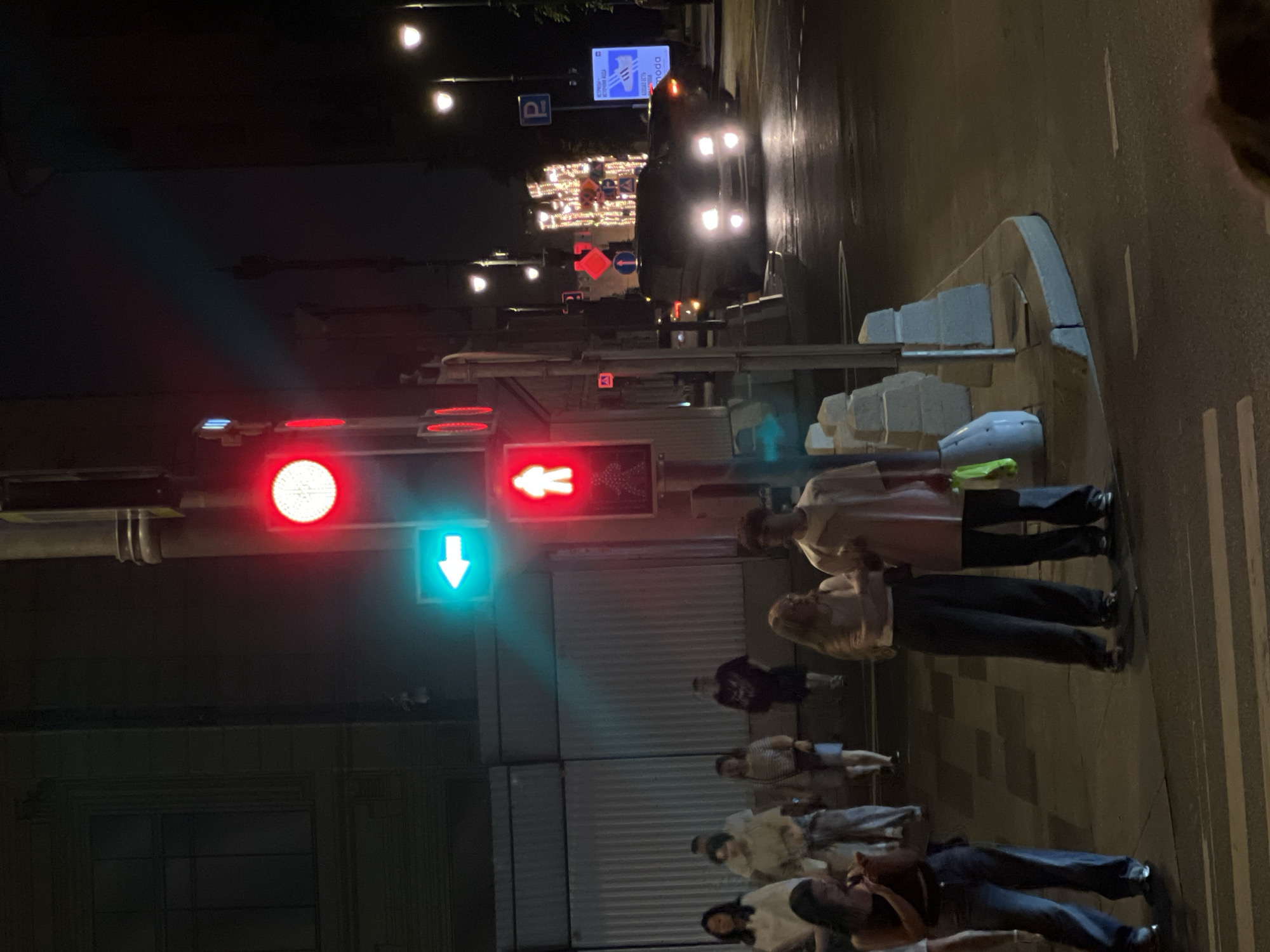 In a nighttime urban scene, a traffic light displays a red illuminated figure and a green left-pointing arrow. Several people are visible on the sidewalk, some standing and others walking. One person in the foreground wears a dark top and blue jeans. To their right, a couple stands together, the woman in dark pants and a light-colored top, and the man in a light-colored long-sleeved shirt and dark pants. Further back, more individuals are present, some obscured by distance or other people. Across the street, a dark-colored van with its headlights on is positioned at an intersection, suggesting traffic is present. Streetlights and decorative lights are visible in the background, illuminating the urban environment. The ground is paved with tiles and asphalt, with visible crosswalk markings. The scene appears to be a street corner in a city.