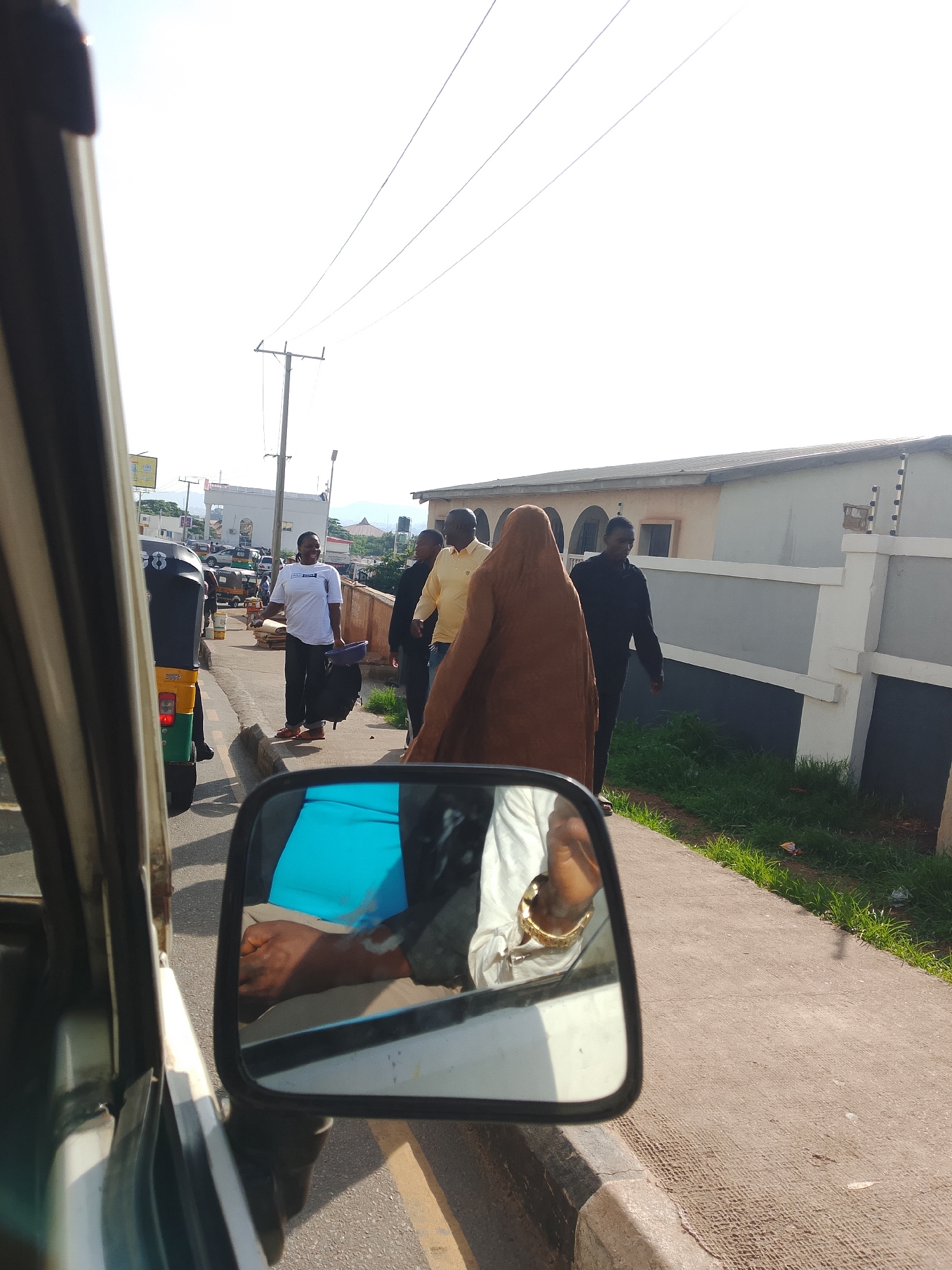 The image captures a street scene from inside a vehicle, likely a car or van, as evidenced by the wing mirror and part of the interior in the foreground. The mirror reflects a person with a hand adorned with a gold bracelet, wearing a white garment. Outside the vehicle, several individuals are visible on a paved sidewalk next to a road. One person in the foreground of the sidewalk is wearing a white t-shirt and dark pants, holding a black bag and a purple basket, and appears to be smiling. Further down the sidewalk, a person in a brown robe is walking, followed by a person in a black long-sleeved shirt. Another individual in a yellow long-sleeved shirt is also present. A few meters away, another man in a white t-shirt is seen carrying bags. In the background, there is traffic, including a three-wheeled auto-rickshaw, and buildings lining the street. Utility poles with wires are also visible against a bright sky. The environment appears to be a sunny day in a built-up area.