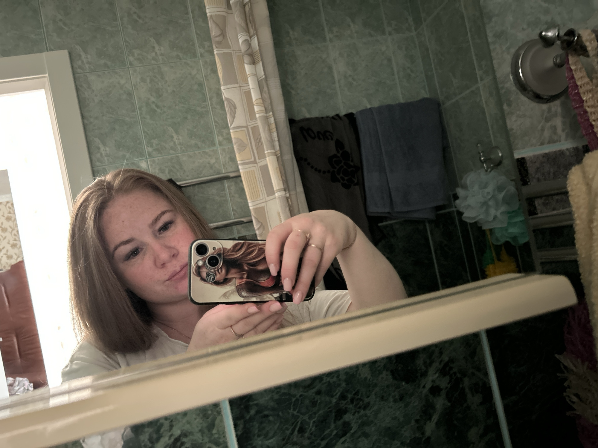 The image shows a young woman taking a selfie in a bathroom. The location is Chaikovsky, Russia.
The woman has light brown hair and is wearing a white shirt. She is holding a smartphone with a case featuring an image of a woman. The bathroom has green tiled walls and a mirror. A shower curtain, towels, and bathroom accessories are visible in the background.