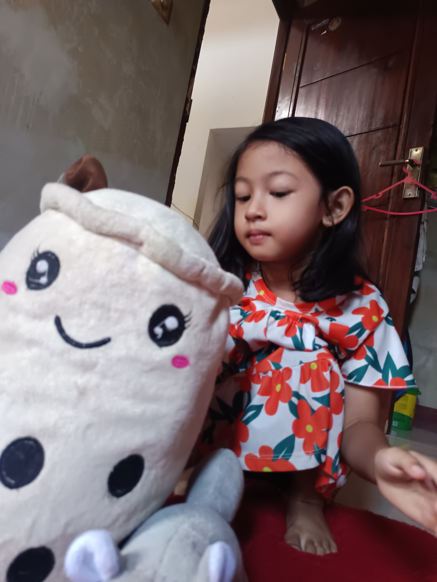 In an indoor setting, likely a home in Maleber, Indonesia, a young girl with dark hair is seated on a red surface, possibly a rug. She is wearing a white dress adorned with a vibrant floral pattern featuring orange flowers and green leaves. To her left, a large, plush toy resembling a smiling cup of bubble tea is prominently displayed. The toy is light-colored with black circles for eyes and a curved line for a mouth, along with pink blush marks on its cheeks. Partially visible in front of the bubble tea toy is another smaller gray stuffed animal. The background includes a textured, light-colored wall and a dark wooden door with a brass handle and a pink hanger. The lighting suggests an indoor environment, possibly during the daytime.