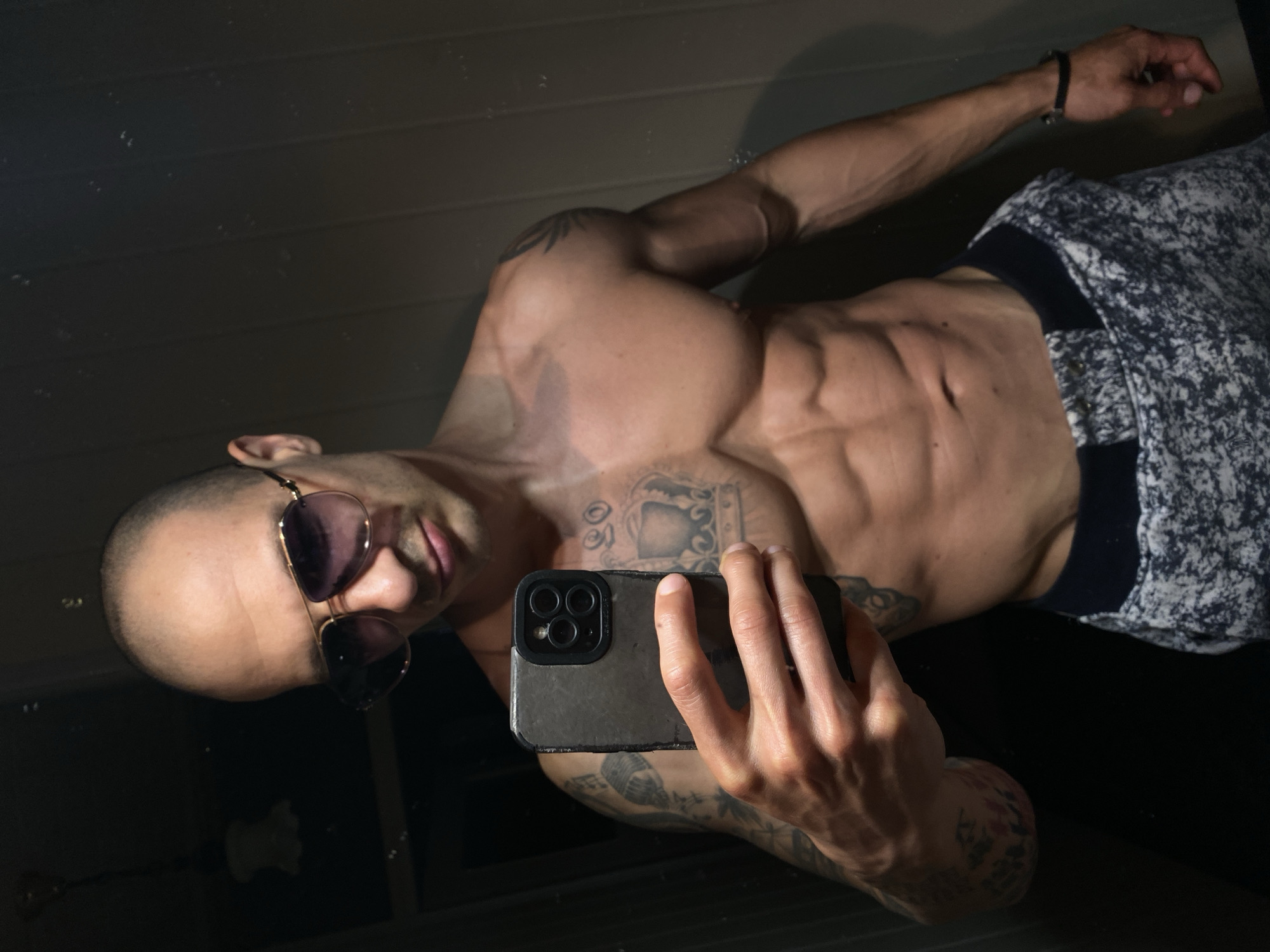 A shirtless man with sunglasses is taking a selfie in a dark room. He has tattoos on his chest, arms, and shoulder. He is wearing a patterned shirt and a black bracelet. The room appears to be a bathroom, with a dark, reflective surface behind him. There is no information available to determine if this photo was taken in Pleven, Bulgaria.