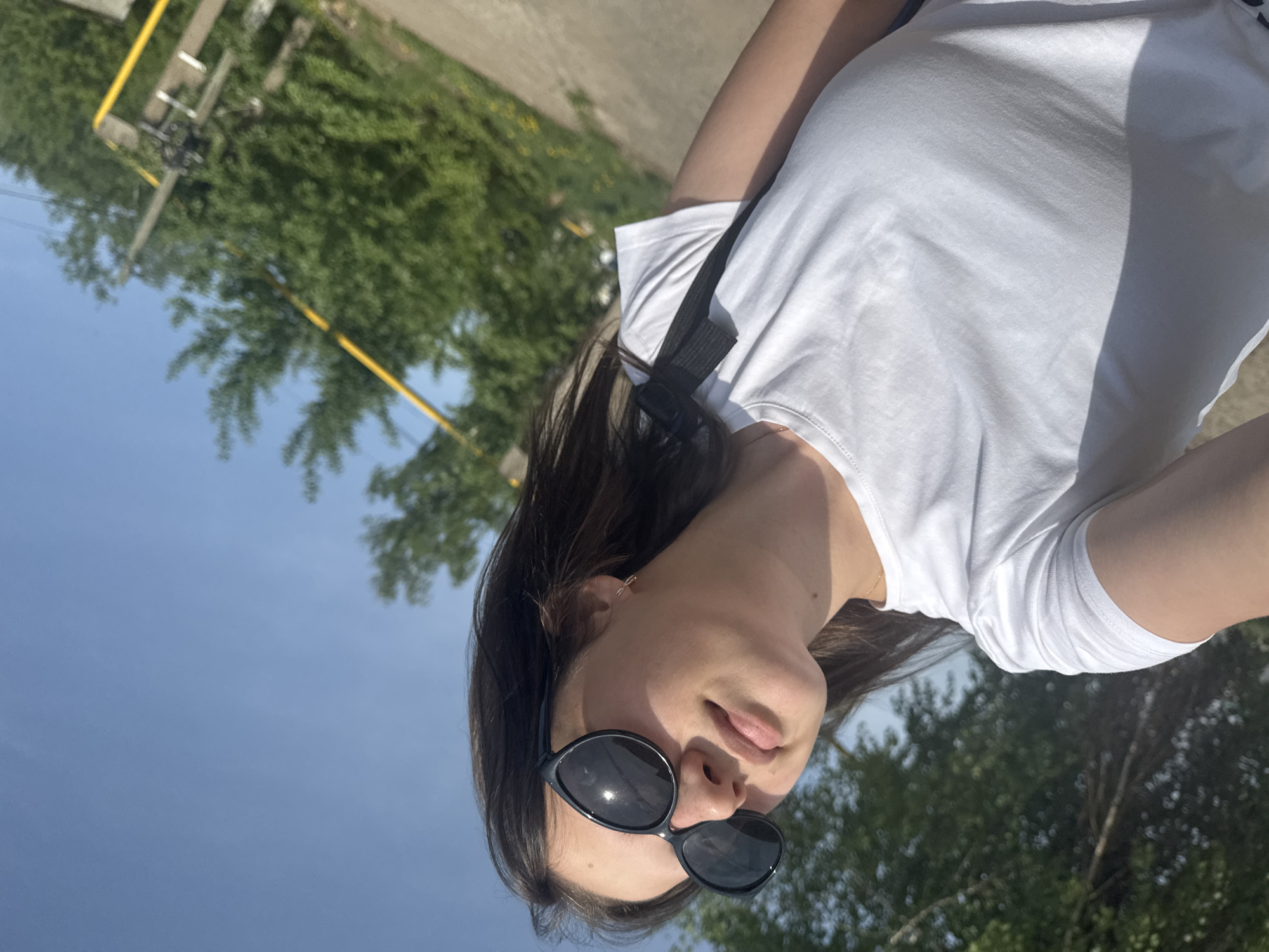 A woman is pictured from a low angle, presumably taking a selfie outdoors. She is wearing sunglasses and a white T-shirt, with a black strap visible across her shoulder. The background includes trees, a blue sky, and a yellow metal structure that may be related to infrastructure. The image appears to be rotated counter-clockwise. The location is Chaikovsky, Russia.