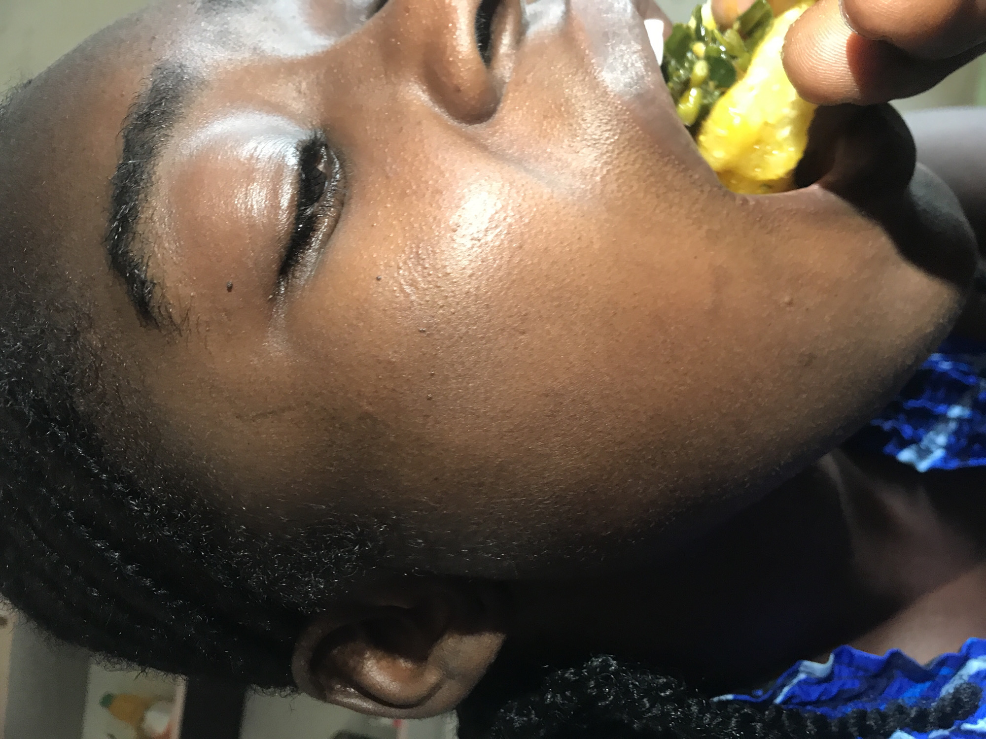 A close-up, profile shot shows a person in New Stadium, Nigeria, eating a piece of food. The person's head is turned to the right, with their left eye closed and their mouth open to receive the food. The food appears to be yellow and green, possibly a fruit with vegetables. The person has dark skin and braided hair, and is wearing a blue and white patterned garment. The background is blurred, but appears to be indoors.