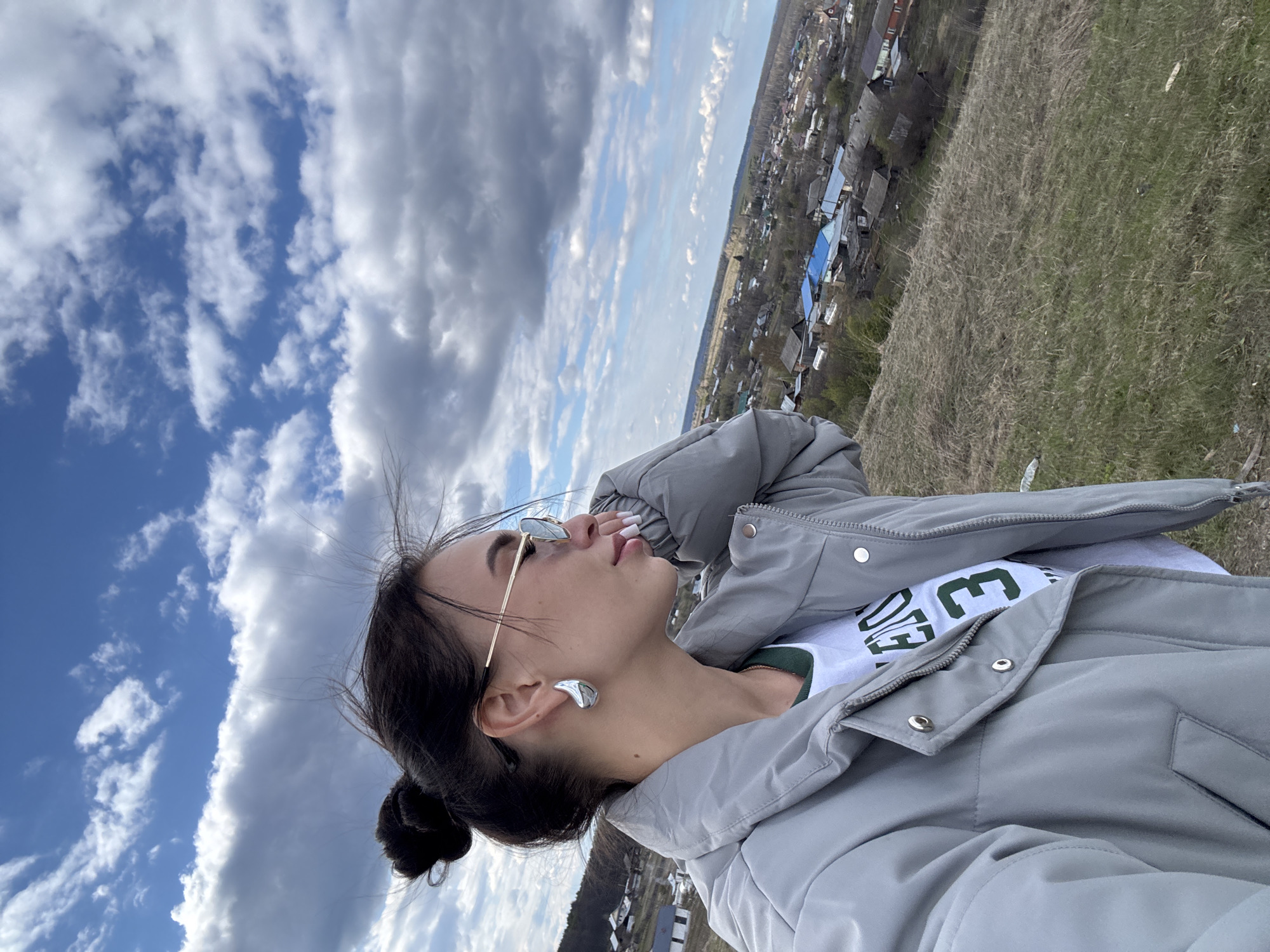 The image, rotated 90 degrees clockwise, captures a young woman posing outdoors on a bright, partly cloudy day. She is shown from the chest up, looking upwards and away from the camera with a serene or contemplative expression. Her dark hair is pulled back in a low bun, with some strands appearing to be gently moved by the wind. She wears slim, gold-framed sunglasses and prominent silver-toned, organically shaped earrings. Her clothing consists of a light gray puffer jacket worn over a white t-shirt. Green lettering is visible on the t-shirt, showing parts of a word like "OVE" and a separate "E".
The background features a expansive sky with patches of bright blue interspersed with large, fluffy white and gray clouds. Below the horizon, a distant settlement spreads across a wide landscape, revealing numerous houses with various roof colors, including distinct bright blue sections. The immediate foreground shows a natural hillside covered with dry, light brown and green grass. The scene suggests an elevated viewpoint over a rural or suburban area, characteristic of a location like Фоки, Russia.