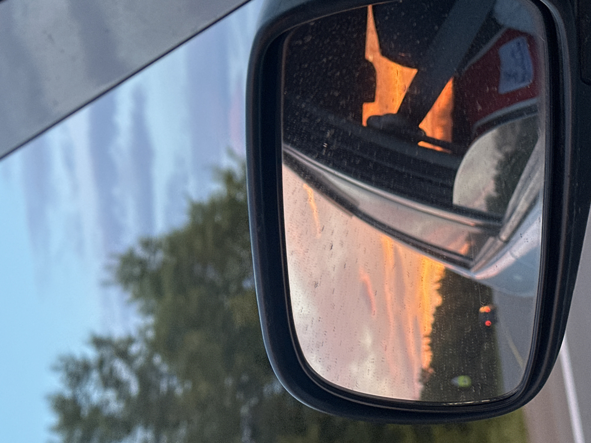 The image shows a car's side mirror reflecting a sunset. The mirror is framed by a black housing and shows a vibrant orange and pink sky with some clouds. In the reflection, trees are visible along the horizon, and the lights of another vehicle are seen on a road. The interior of the car is partially visible, including a steering wheel and a seat. The exterior of the car is a light silver color, and the sky above is a pale blue with streaks of grey clouds. There are visible water spots on the mirror. The scene suggests travel, possibly on a highway outside of a city. The image does not contain enough information to determine the location in Yelizovo, Russia.