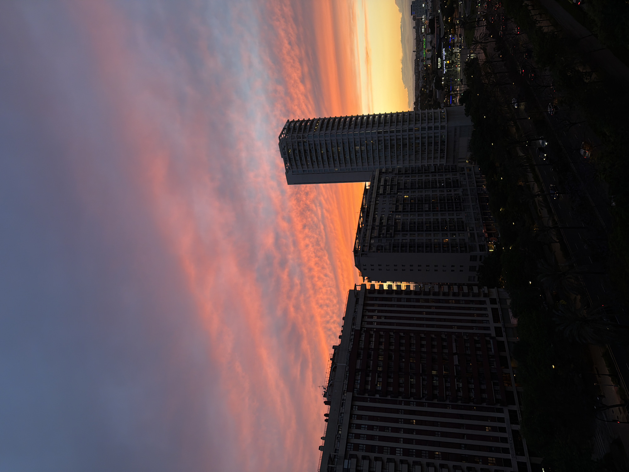 The image shows a cityscape at dusk in València, Spain. The sky exhibits a gradient of colors, from deep blue to vivid pink and orange, with streaks of cirrus clouds that add texture. Three high-rise buildings dominate the foreground. They are constructed with geometric lines and rectangular windows. The city's skyline is visible in the distance, marked by a mix of illuminated structures and trees. A main road runs along the right side, lined with trees and interspersed with lights from vehicles.