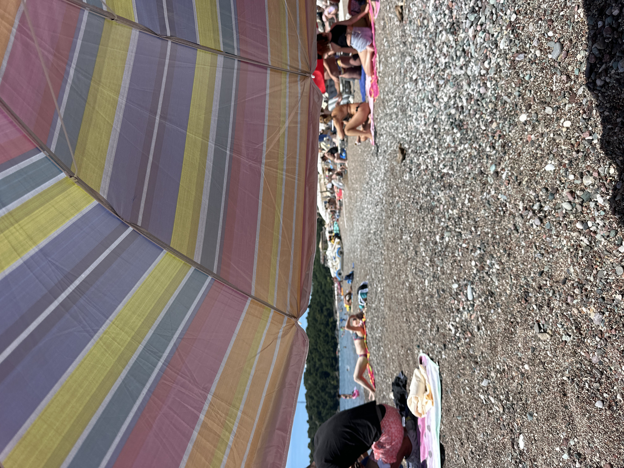 The image depicts a crowded beach scene in Boreti, Montenegro, on a sunny day. A large, colorful striped umbrella dominates the foreground, casting a shadow over the pebbled shore and several beachgoers. In the mid-ground, a woman in a bikini lies on a towel, facing the viewer and propped up on her elbows, with a blurred green forested hillside and the Adriatic Sea in the background. Other individuals are scattered on towels and lounge chairs, some in swimsuits, others dressed in casual beachwear. Boats are visible in the water. The beach is covered in smooth, grey and multi-colored pebbles.
