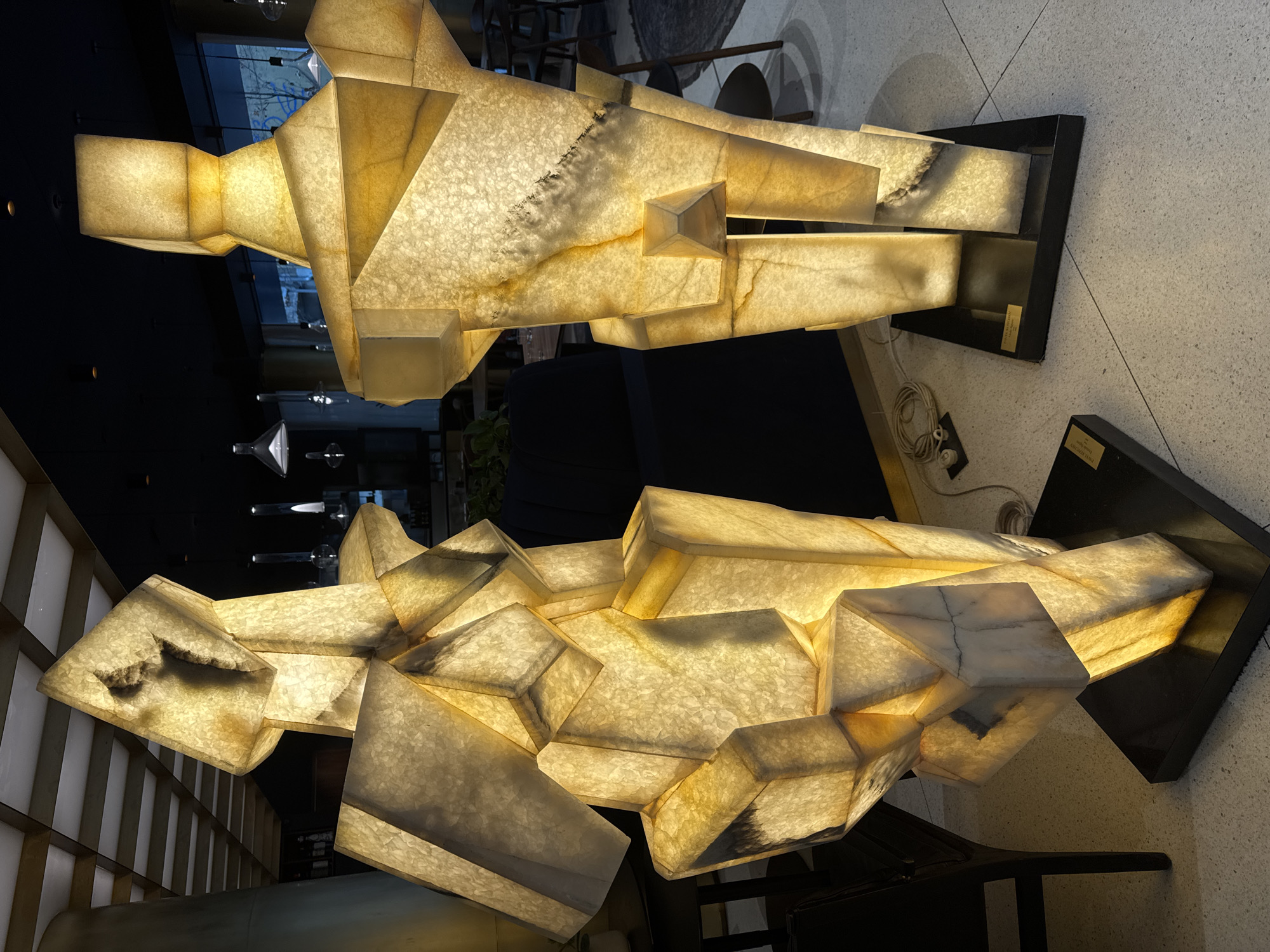 Two large, internally illuminated, anthropomorphic sculptures stand on a light-colored terrazzo floor within a modern interior space in Sofia, Bulgaria. Each sculpture is composed of numerous translucent, yellowish-white geometric segments, resembling faceted human figures and exhibiting natural veining and darker inclusions consistent with onyx or a similar stone. They emit a warm glow and are mounted on dark, rectangular bases.
The sculpture on the left is positioned slightly forward and is composed of more angular, stacked blocks. The sculpture on the right stands taller and has a more defined, upright posture with a distinct pyramidal element at its lower torso.
The background is dimly lit, featuring dark furniture including chairs and tables, indicative of a hotel lobby or upscale establishment. Large windows reveal a blurred urban outdoor scene. Overhead, the ceiling is dark with visible recessed lighting fixtures and a section of a white grid ceiling structure on the upper left. White coiled electrical cables are visible on the floor near the base of the right sculpture. No people or active interactions are present in the scene.