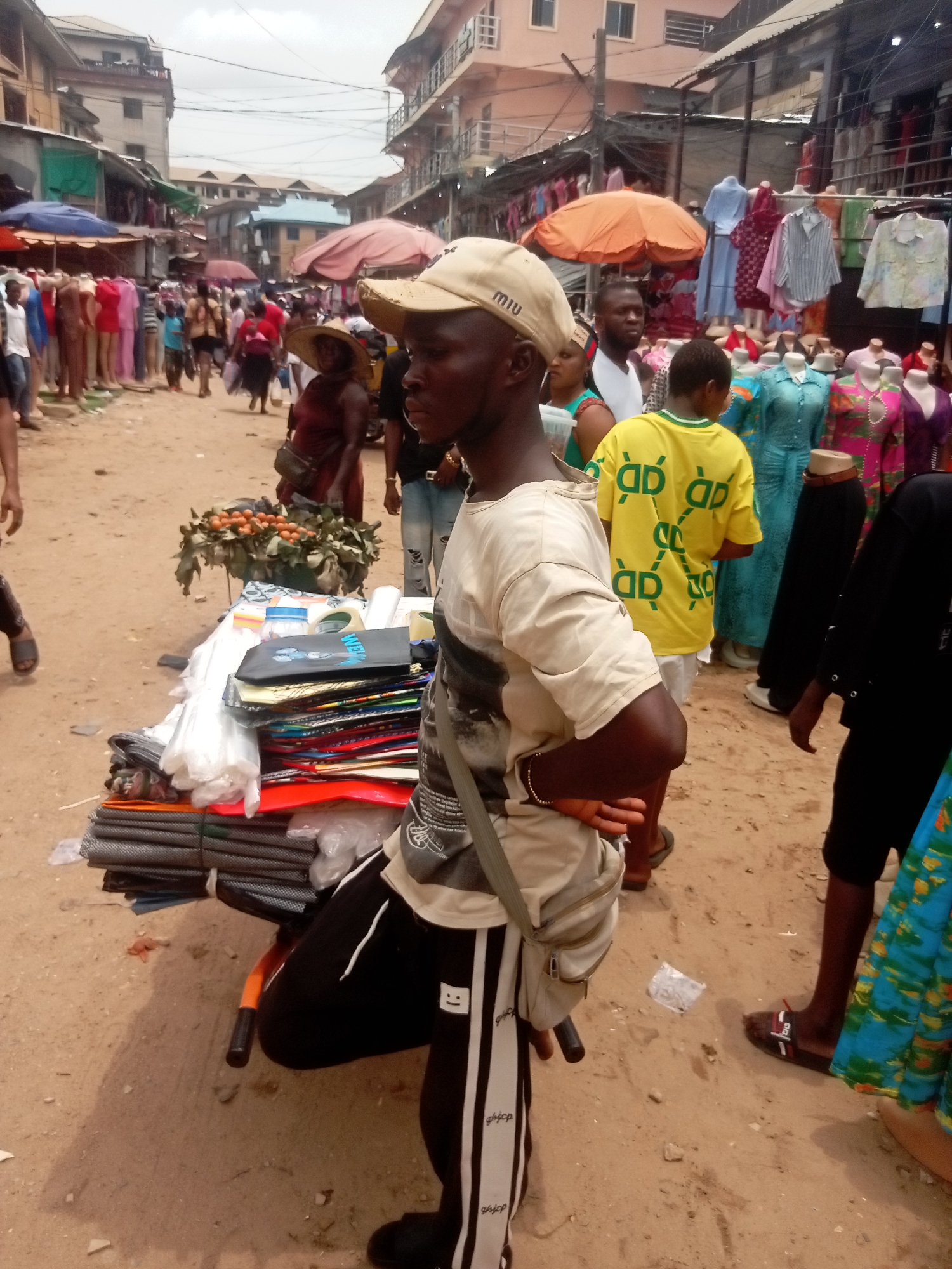 The image depicts a crowded market scene in Fegge, Nigeria. The ground is sandy. A man in the foreground, wearing a beige cap, a white t-shirt, and black-and-white striped pants, is leaning on a small cart loaded with various items, including fabric, folders, and a basket of oranges. He has a bag slung across his shoulder. Behind him, there are numerous people, some standing, some walking. Clothes are displayed for sale under orange and blue umbrellas. Buildings line the sides of the street, and overhead, there are numerous wires. The lighting suggests an overcast day.