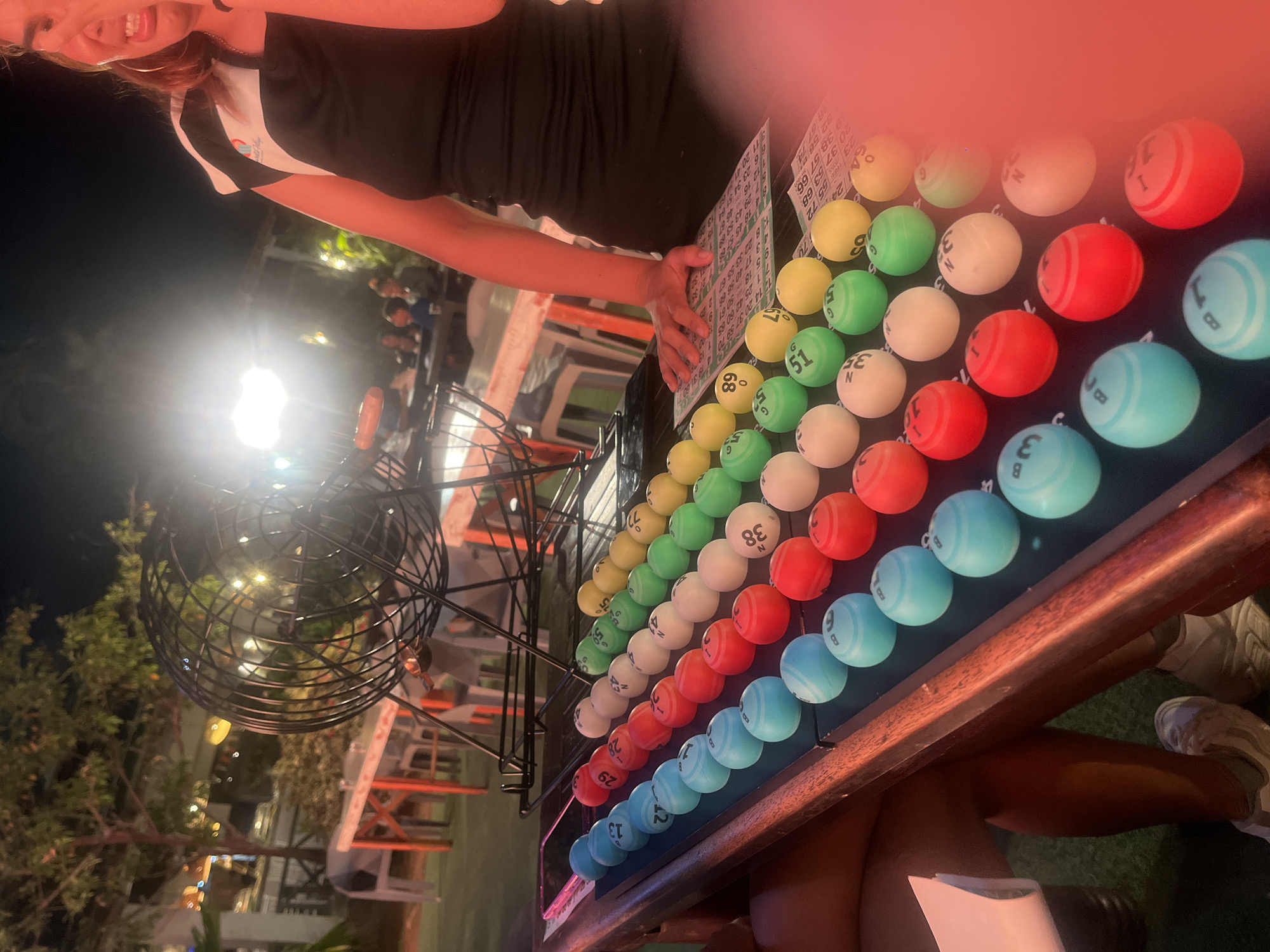 In Porlamar, Venezuela, a woman is seen standing next to a table displaying a bingo game. The scene is outdoors, possibly at night, with ambient lighting from nearby establishments. The woman, partially visible, is wearing a black dress with white accents on the shoulder area and large hoop earrings. Her facial expression is one of distress or discomfort.
In front of her is a table set up for bingo. A large metal cage, typical for bingo ball dispensing, is positioned behind the table. The table itself is covered with rows of colored balls, arranged in columns. The balls are blue, red, white, green, and yellow, and some have numbers printed on them, such as "13", "27", "29", "38", "49", "51", "65", and "89". Bingo cards are also visible on the table, with some numbers marked. The table has a dark surface and a wooden edge.
The background suggests an outdoor venue with tables and chairs, likely a restaurant or entertainment area. There are trees with lights in them, and other patrons are visible in the distance, seated at tables. The overall atmosphere appears to be social and recreational.