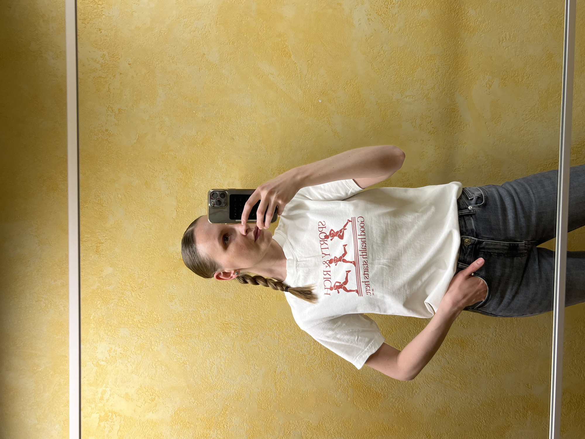 An indoor mirror selfie captures a young woman standing in what appears to be a room with a textured yellow wall, in Khimki, Russia. The woman is reflected in a mirror, whose thin white frame is visible along the left and right edges of the image. She is wearing a white short-sleeved t-shirt and dark grey jeans. Her light brown hair is styled neatly, pulled back from her face, and gathered into a single braid that rests over her left shoulder. She is holding a dark-cased smartphone in her right hand, using it to take the photo, while her left hand is casually tucked into the pocket of her jeans. She has a neutral expression, looking towards the phone screen. On her t-shirt, visible text in red, appearing backward due to the mirror reflection, reads "GOOD HEALTH IS GOOD WEALTH" and "SPORTY & RICH." The t-shirt also features three stylized figures in active, running poses. She wears a small stud earring in her left ear and a ring on the ring finger of her right hand. The lighting suggests it is daytime.