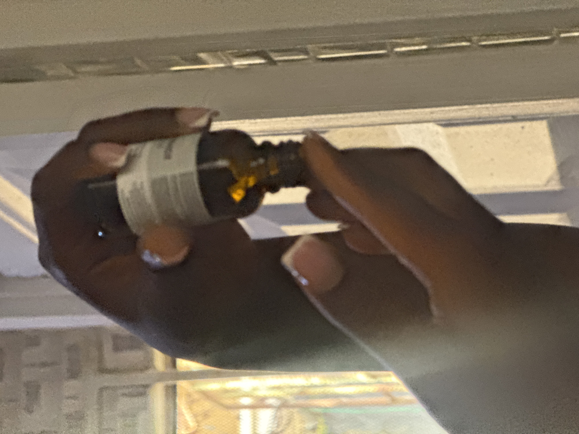 The media file shows a close-up of two hands holding a small, dark glass bottle with a dropper. The hands appear to belong to a person with dark skin tone and manicured nails. The bottle has a white label with some text, though it is mostly illegible due to the resolution. A small amount of amber-colored liquid is visible inside the bottle, and some of it has dripped onto the dropper. The background is blurred, but it seems to be an indoor setting with some artificial lighting. The overall lighting suggests it could be daytime or evening. The context provided is Abuja, Nigeria.