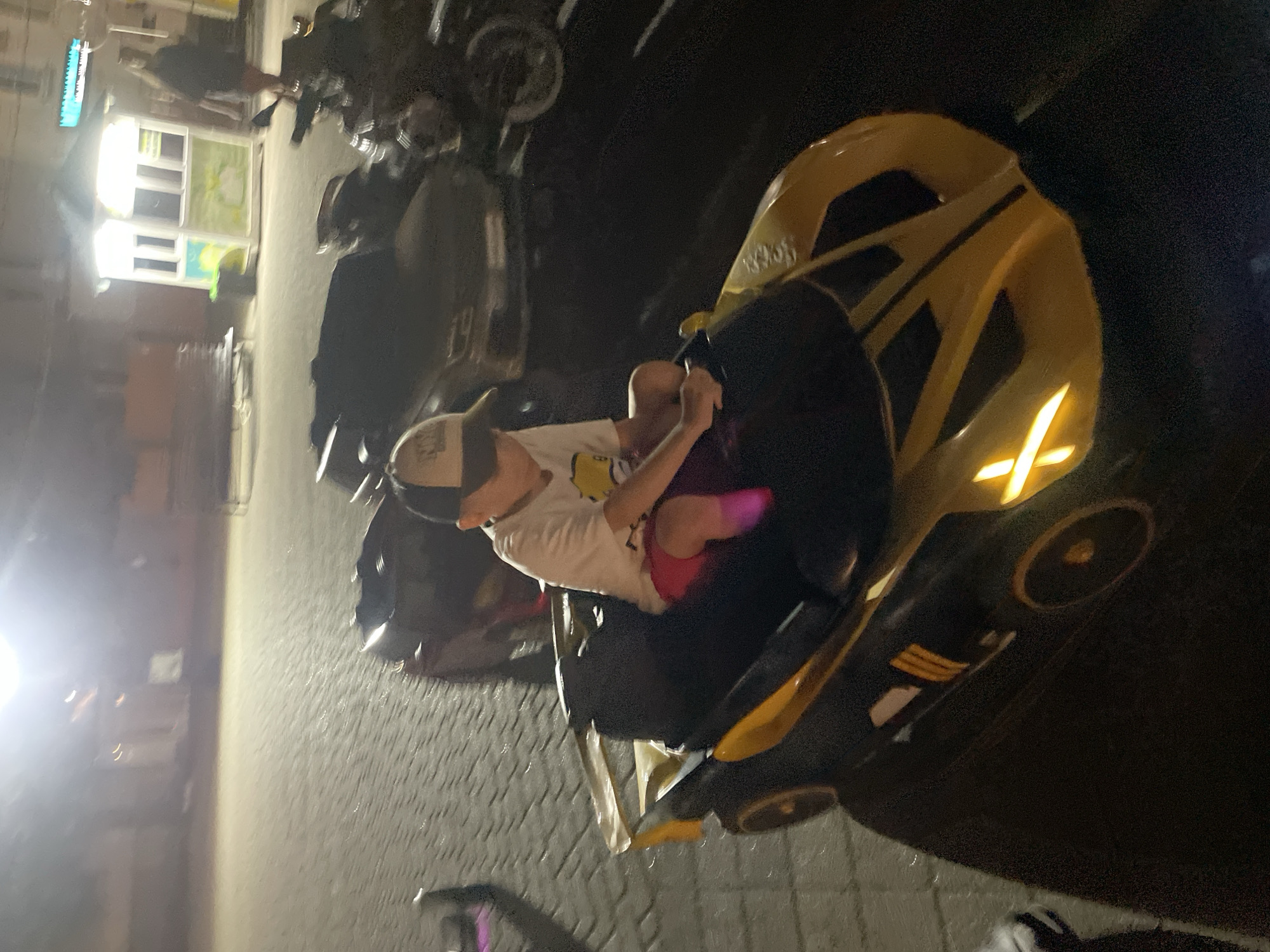 The image shows a child wearing a hat and a t-shirt, sitting in a yellow and black toy car. The child is holding onto a steering wheel. In the background, there are other toy cars, a motorcycle, a small building with illuminated windows, and a paved area. The lighting suggests it is nighttime or evening. The scene appears to be an outdoor recreational area in the city of Merchanskoe, Russia.