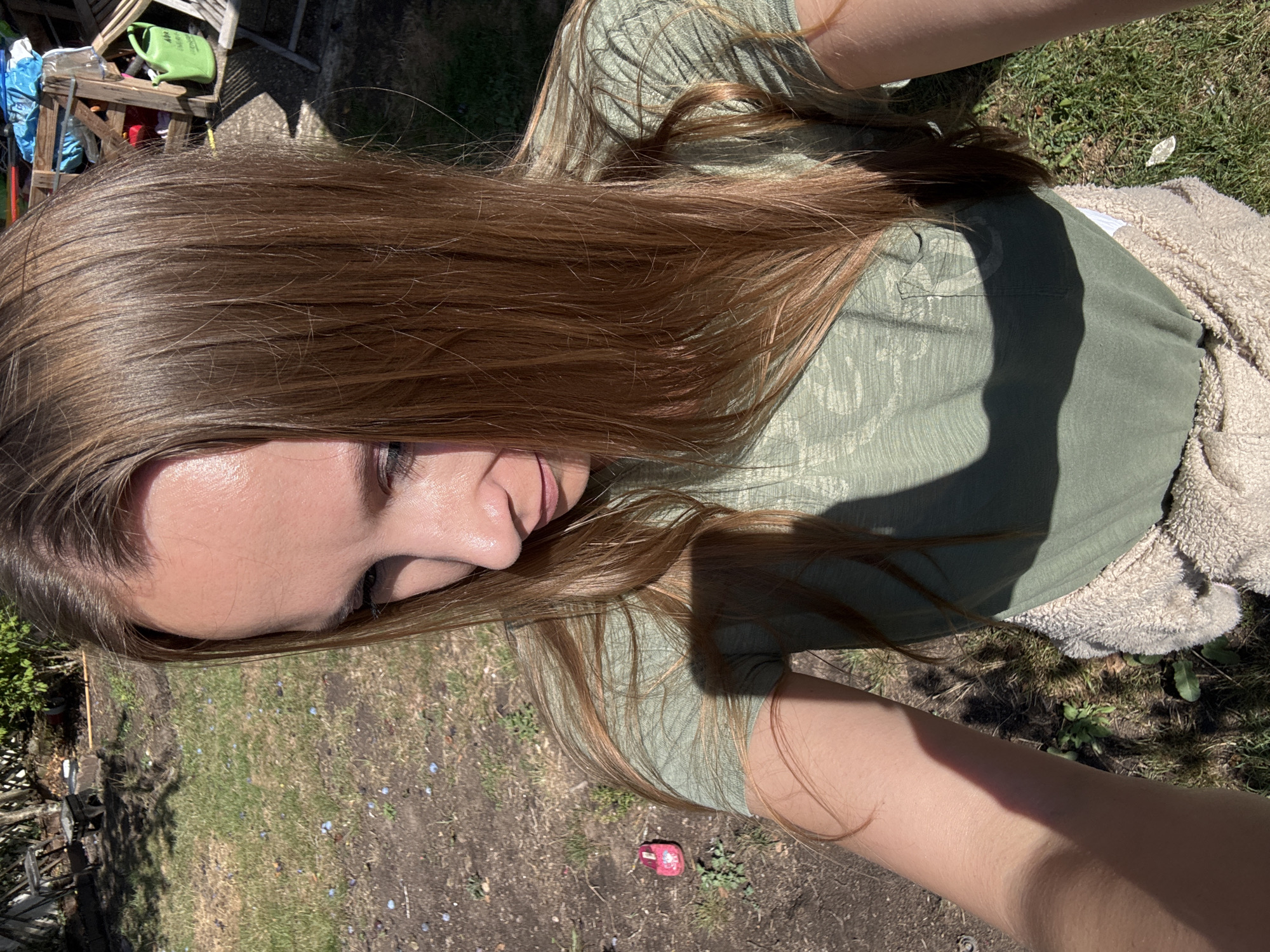 A woman is taking a selfie outdoors in a backyard environment. She has long, brown hair that is partially covering her face as she looks down and to her left. She is wearing a green t-shirt and a tan, fuzzy garment tied around her waist. The background shows a grassy area with patches of dirt, some scattered small blue objects, and a small red toy in the lower left quadrant of the image. Further in the background, there is some wooden fencing, debris, and a green watering can. The lighting suggests it is a sunny day. There is no specific identifiable information that places this image in the city of Überherrn, Germany.