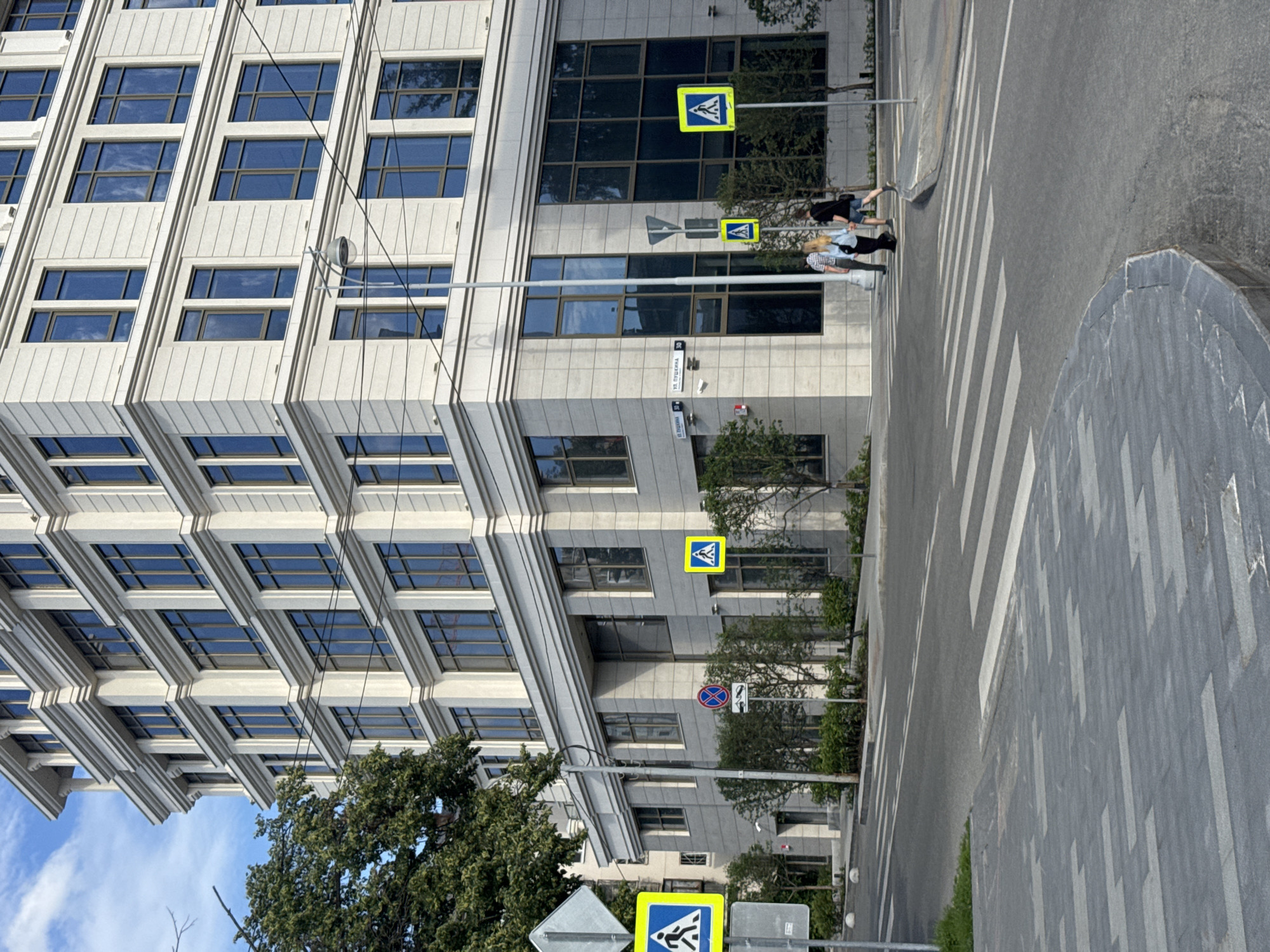 The image shows a street scene in Yekaterinburg, Russia, with a modern office building and a pedestrian crossing. Two people are visible crossing the street at the pedestrian crossing. The weather appears to be sunny, and the time of day is likely daytime, given the bright lighting. The building is made of concrete and glass, with many windows. A street sign is visible with Cyrillic text, and pedestrian crossing signs are also present. The scene is urban and depicts a typical day in a city.