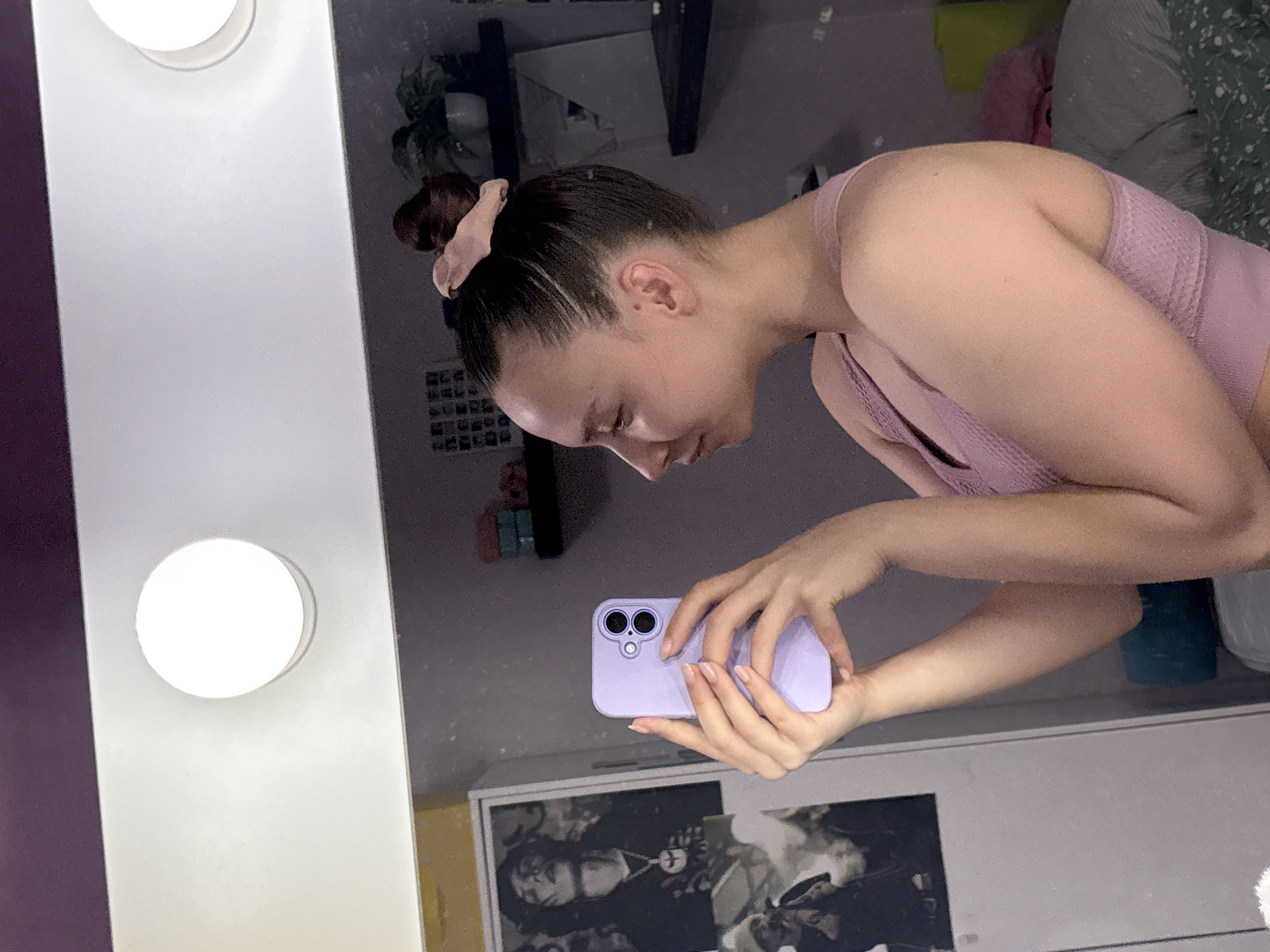 A young woman with dark hair pulled into a bun with a pink scrunchie is looking at a lavender-colored smartphone in her hands. She's wearing a light purple textured tank top. The reflection shows she's in front of a lighted vanity mirror. The background features shelves with plants, artwork on the wall, and a partially visible bed. Based on the prompt, this scene is located in Прикамский, Russia.