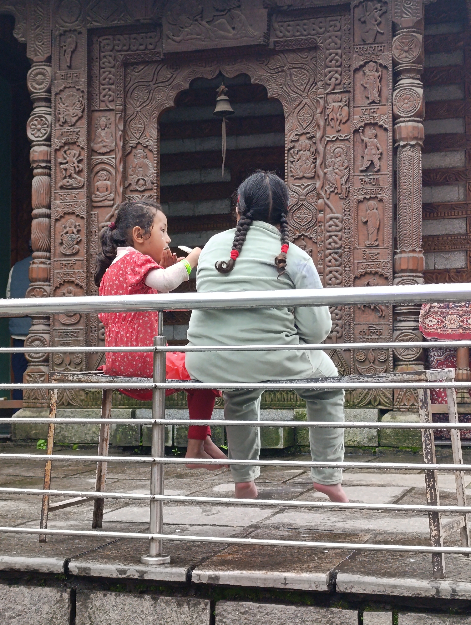 The image depicts two girls sitting on a metal bench in front of a temple in Manali, India. The girl on the left, facing away from the camera, is wearing a red dress with white polka dots and has her hair in a ponytail. She appears to be holding something white up to her mouth. The girl on the right, also facing away, is wearing a light green top and pants, and her hair is in two braids with red ribbons. The temple behind them features intricate wooden carvings depicting various figures and symbols, characteristic of Himalayan architecture. The ground is paved with stone tiles, and there are metal railings in the foreground. The scene suggests a moment of rest or observation in a culturally significant location.