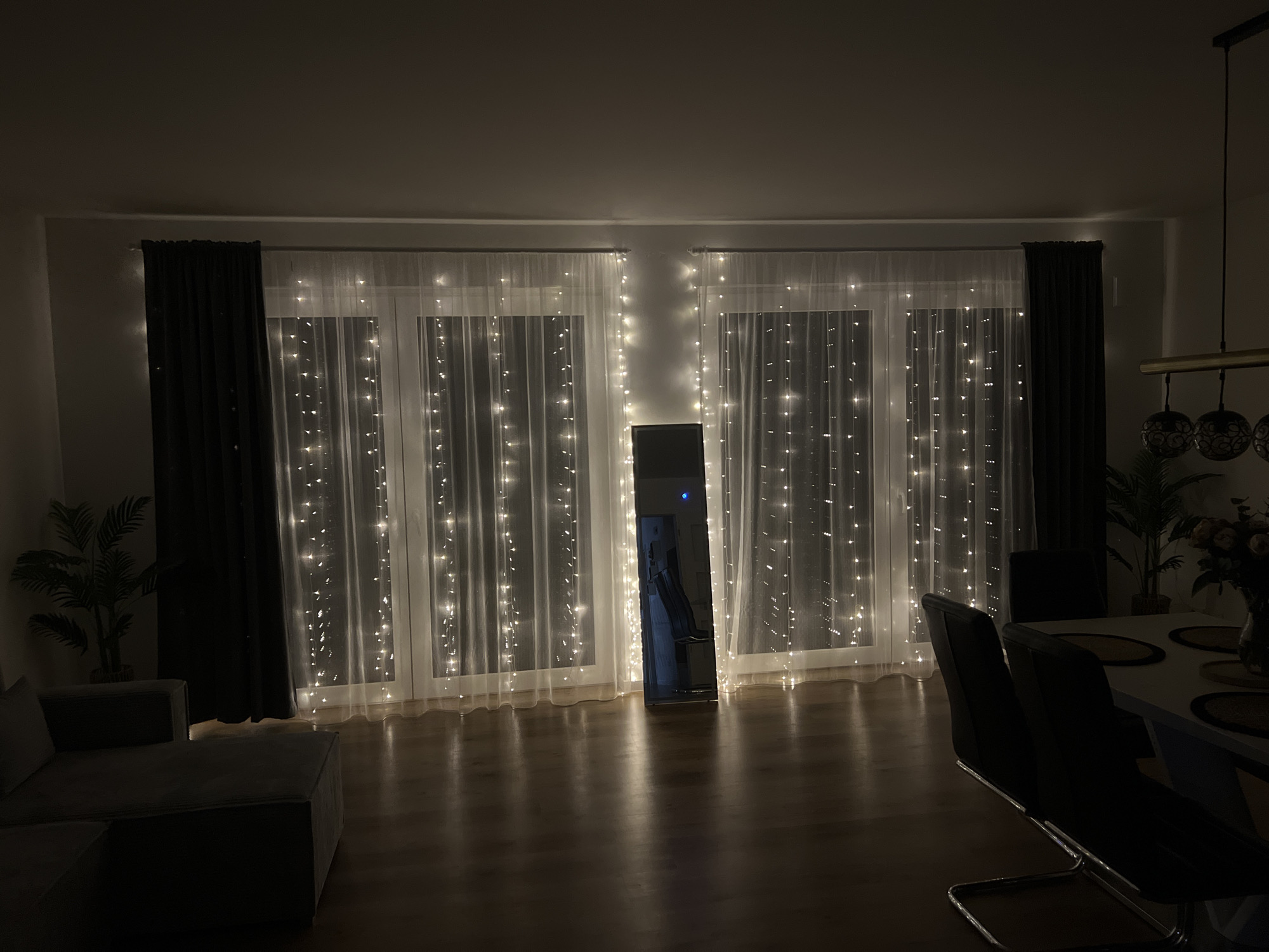 This is a low-light, wide-angle interior shot of a living and dining area, likely in Bad Zwischenahn, Germany. The scene is illuminated by numerous string lights draped across two large windows, creating a festive and warm ambiance. A dark grey sectional sofa is positioned to the left of the frame, partially obscuring the left window. To the right, a dining table with four black chairs is visible, accompanied by a vase of flowers. A tall, full-length mirror stands between the two windows, reflecting the string lights and a glimpse of another room. The floor is made of polished wood, reflecting the light.
