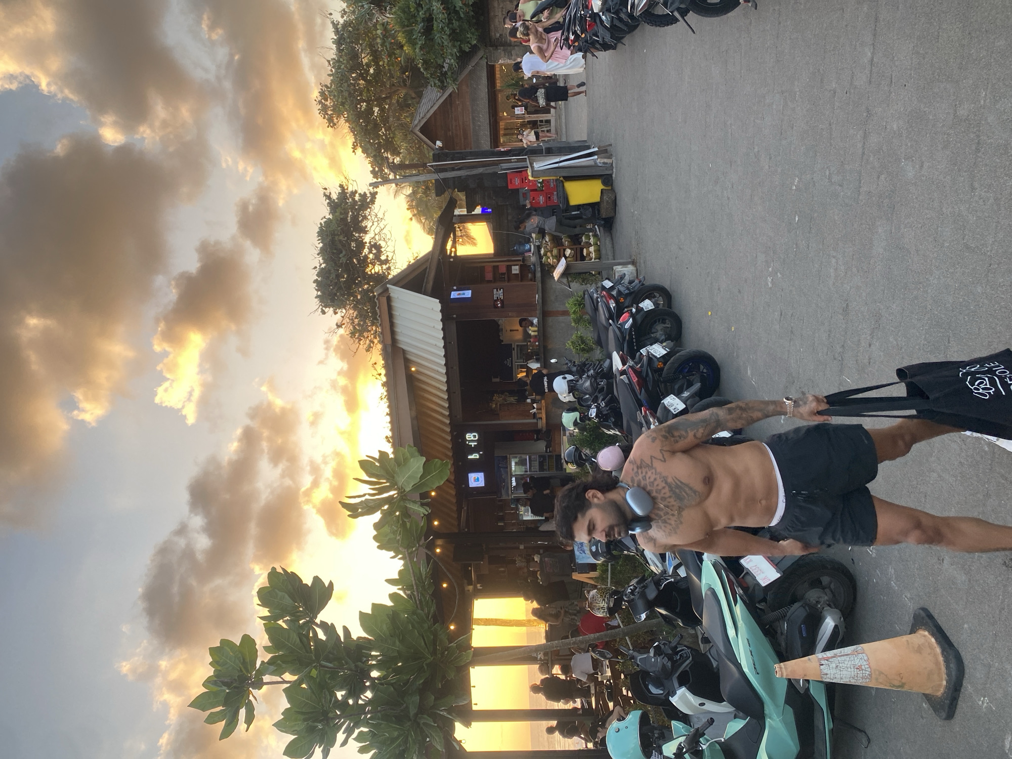 The image depicts a street scene in North Kuta, Indonesia, at sunset. In the foreground, a shirtless man with tattoos and headphones around his neck walks towards the right of the frame, carrying a black tote bag. He is wearing black shorts. Behind him, numerous motorcycles are parked. To his left, a turquoise scooter is visible. In the background, several open-air establishments with awnings line the street, with people dining or socializing. The sky above is filled with dramatic clouds illuminated by the setting sun, casting a warm glow over the scene. The overall atmosphere suggests a lively evening at a coastal location.