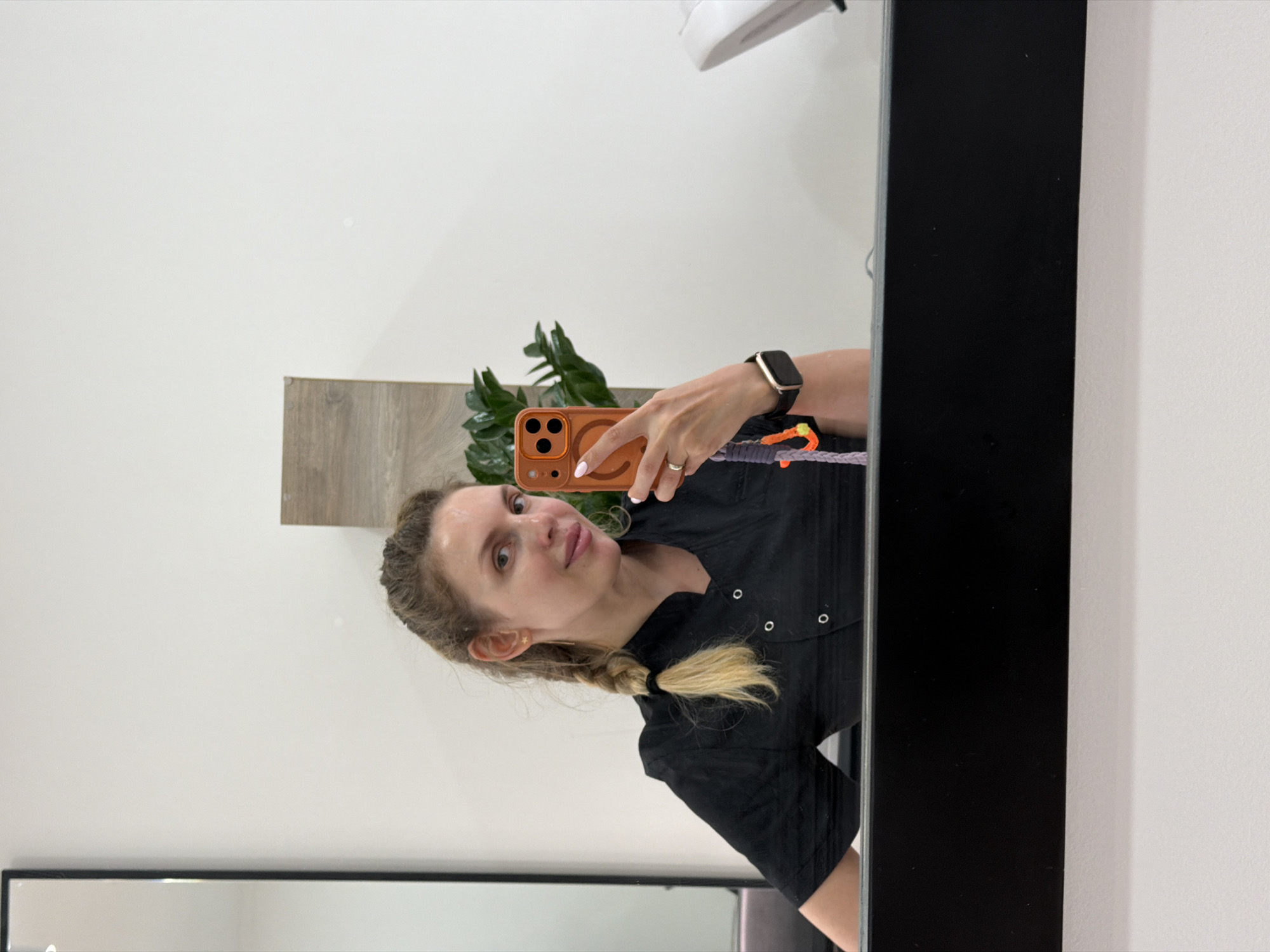 The image, rotated 90 degrees counter-clockwise, captures a woman taking a mirror selfie indoors, likely in Budva, Montenegro. The woman is shown from the chest up, with light brown hair styled in a braid that ends in a blonde ponytail. She is wearing a black short-sleeved top with snap buttons and has a light complexion. She looks towards the camera with a neutral expression and a slight smile.
In her right hand, she holds an orange-colored smartphone, which has a distinct circular magnetic ring (MagSafe-like) on its back and a prominent camera module. A colorful woven strap, incorporating shades of orange, purple, and black, is attached to the phone. On her left wrist, she wears a black smartwatch with a gold or bronze bezel. A simple silver-toned ring is visible on the fourth finger of her right hand, and her fingernails are painted with light pink polish. A small gold-colored star earring is visible on her left earlobe.
The background, reflected in the mirror, features a plain white wall. To the left, a light-colored wooden panel or shelf is partially visible, behind which a green plant with large leaves adds natural decor. A white light fixture is mounted on the wall above. The mirror itself has a thin black frame, visible along the right side of the image. The setting appears to be a brightly lit indoor space. There is no discernible text visible in the image.