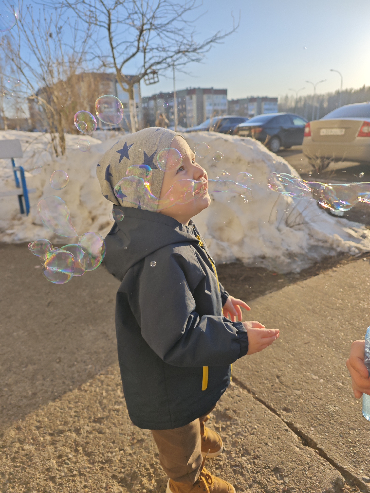 A young child wearing a grey hat with blue stars and a dark blue jacket is blowing bubbles in Markovskii, Russia. The child is standing on a concrete surface with patches of dirt, near a snowbank. Behind the child are several cars and apartment buildings. The sky is bright and clear.