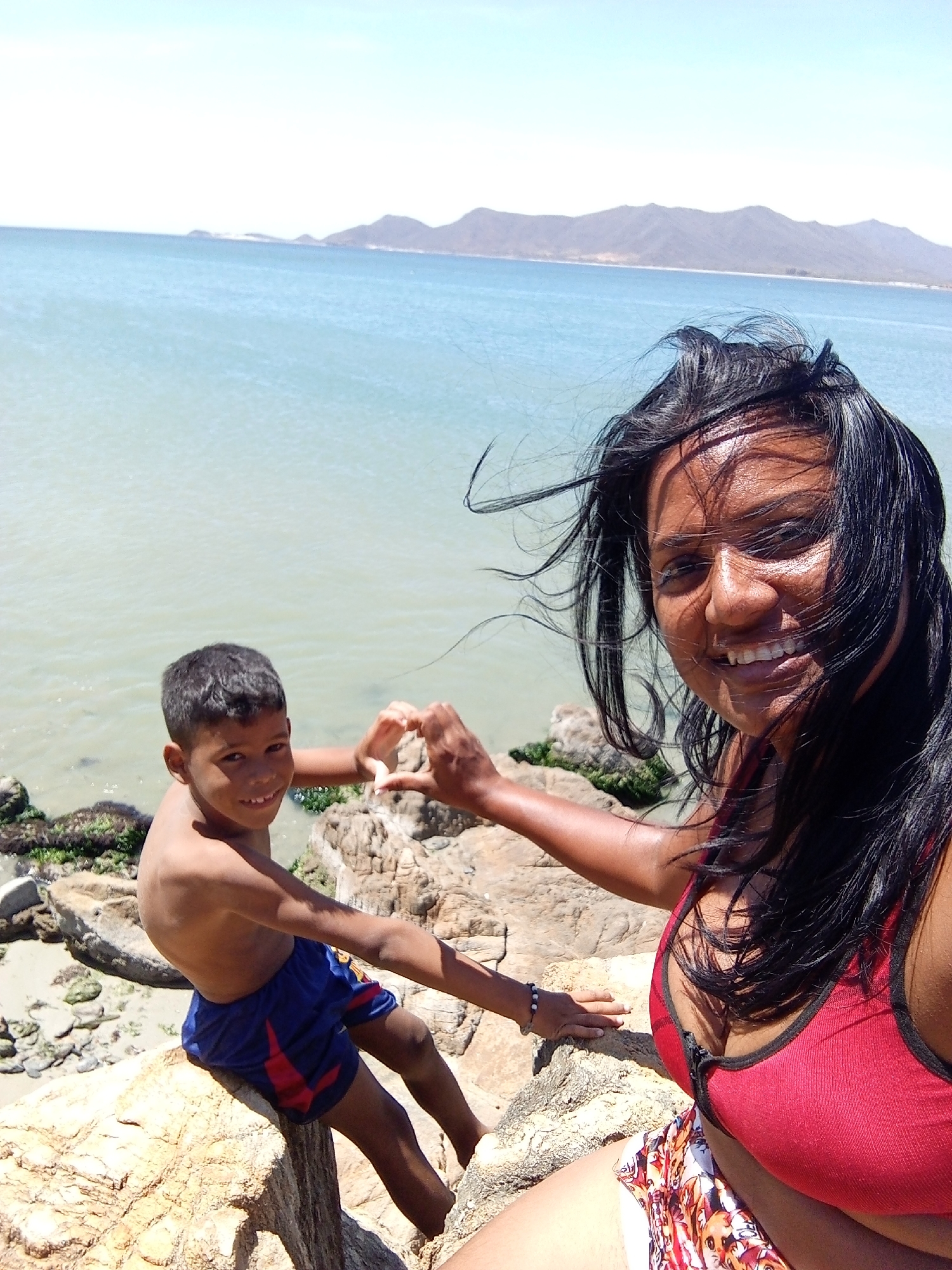The image shows a woman and a boy on a rocky outcrop overlooking a body of water, likely the Caribbean Sea. The woman, with dark hair blowing in the wind, smiles into the camera. She wears a red tank top and patterned bottoms. The boy, bare-chested and wearing blue shorts with red accents, sits on a rock and makes a heart shape with his hands together with the woman. The backdrop consists of the sea and mountainous land on the horizon. The rocks near the edge are covered in patches of green algae or moss. Based on the landscape and the location, it is most likely that this image was taken in or near Juan Griego, Venezuela, given the prompt's context.