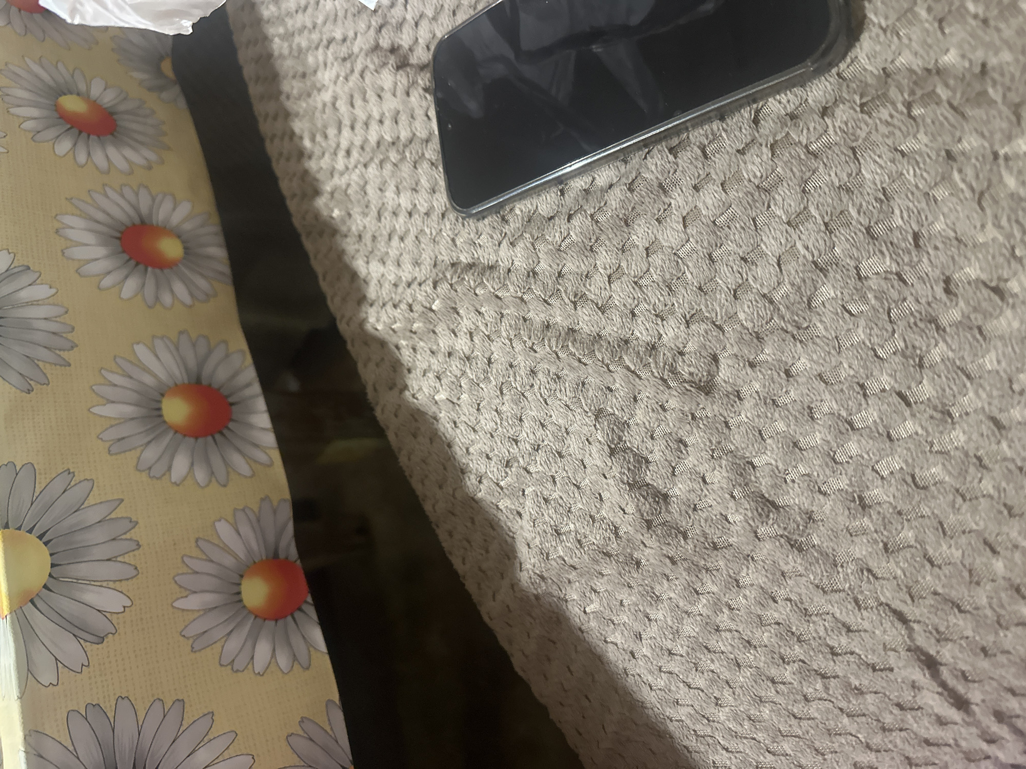 An indoor scene from Razhevo Konare, Bulgaria, displays a static arrangement of household items. A modern smartphone with a dark, inactive screen and a clear or silver-edged case rests on a light brown or grayish-beige textured fabric. This fabric exhibits a distinct repeating pattern of small, raised rectangular or square elements, giving it a waffled or quilted appearance. To the left, separated by a dark, shadowy gap, lies a vibrant yellow fabric adorned with a repeating pattern of large white daisies. Each daisy features a prominent center that transitions from orange to yellow. A crumpled white object, possibly paper or a plastic bag, is visible in the top-left corner, partially obscuring one of the daisies. The lighting suggests an indoor environment, likely under artificial light. No specific activity or event is taking place, and there is no discernible text on any of the visible objects.