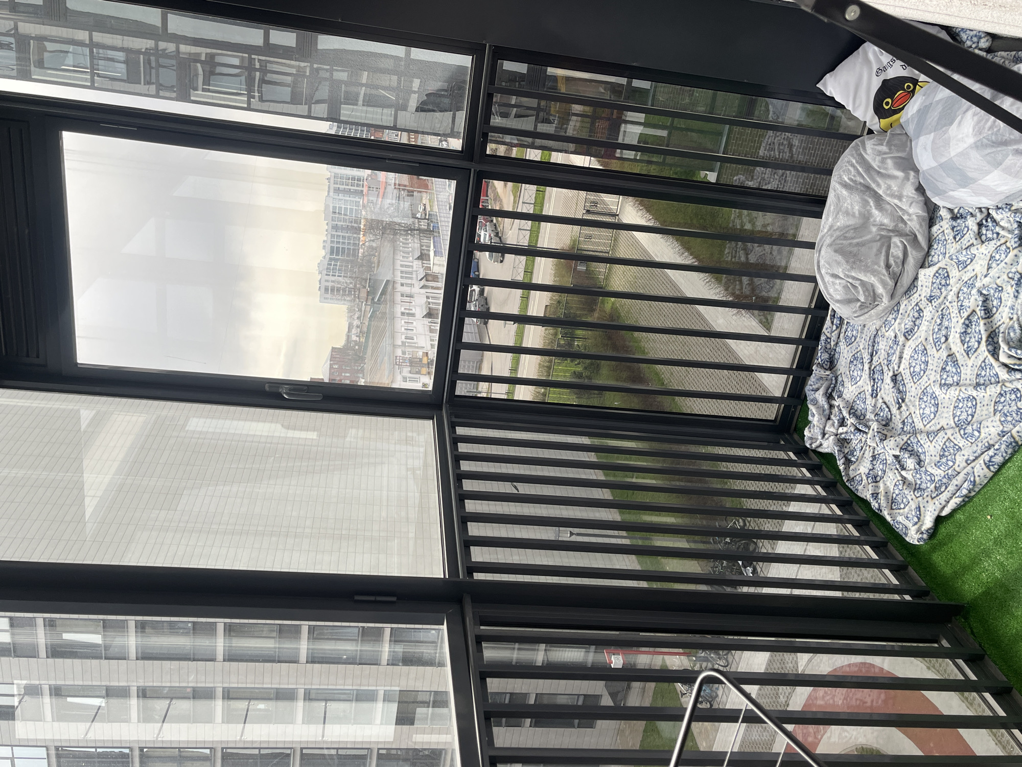 The image shows an elevated view from inside a balcony. Through the glass windows and metal railings, a street scene is visible with buildings, roads, and vegetation. The overall impression is that the location might be a residential building in a city setting, possibly in Saint Petersburg, Russia, as the building style seems typical for Russia. The scene is indoors and on the balcony, with a green area, a blanket and a pillow. No people are visible.