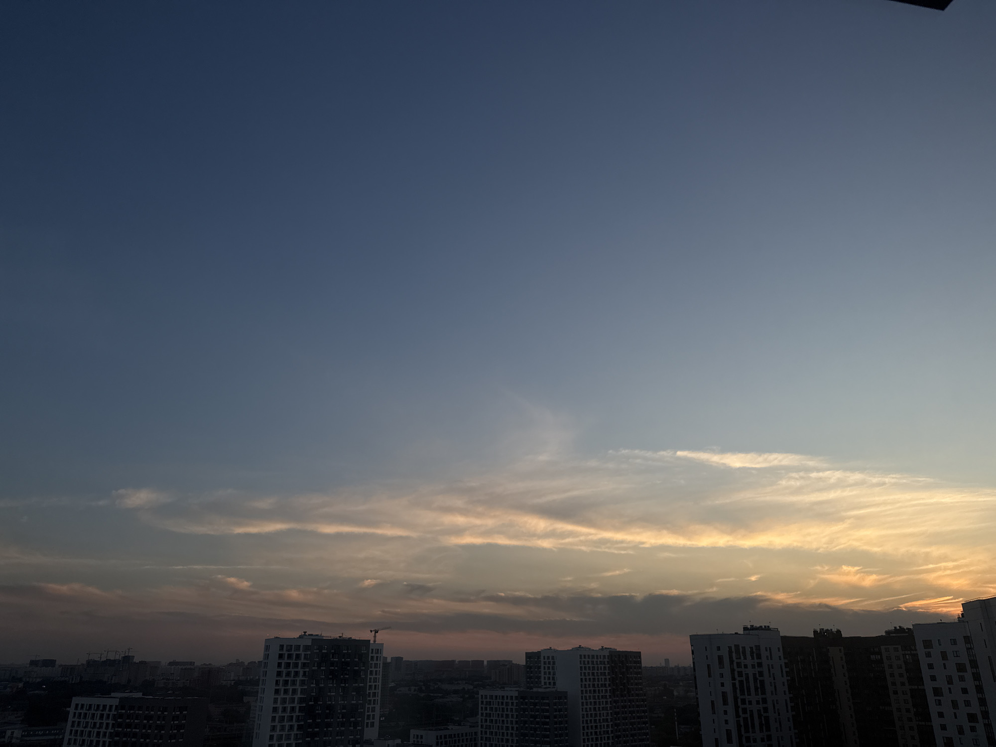 The image depicts a skyline view of the city of Lyubertsy, Russia, at dusk. The sky is a gradient of deep blue at the top, transitioning to lighter blues and then hues of orange, pink, and yellow closer to the horizon, indicative of sunset. Wispy clouds are scattered across the sky, catching the last rays of sunlight. In the foreground, a cluster of modern apartment buildings dominates the lower portion of the frame. These buildings are characterized by their varied heights and contemporary architectural designs, with many windows visible. A few construction cranes are also present in the distance, suggesting ongoing development in the city. The overall scene conveys a sense of urban evening ambiance.