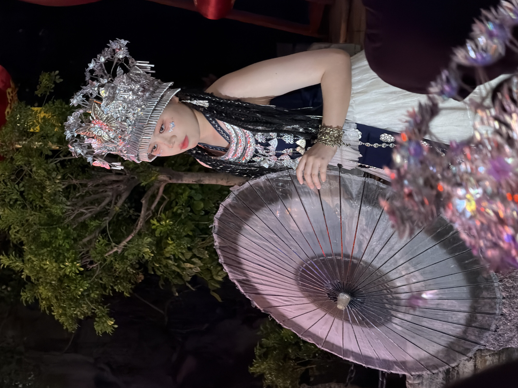 A young woman, adorned in traditional attire with an elaborate silver headdress, is featured in this medium shot. She wears a dark blue top with a white, ruffled skirt, complemented by intricate silver jewelry and bracelets. Her face bears artistic makeup, including subtle pink blush and what appear to be small, sparkly accents above her eyes. The woman holds a large, semi-transparent umbrella in her left hand, its spokes clearly visible. She is positioned outdoors at night, with lush green foliage and a dark, rocky background suggesting a natural or landscaped setting. The lighting is dramatic, with parts of the scene brightly illuminated and others in deep shadow, creating a theatrical atmosphere. The Yuzhong District in China is known for its blend of historical and modern architecture, alongside natural landscapes that could accommodate such a scene.