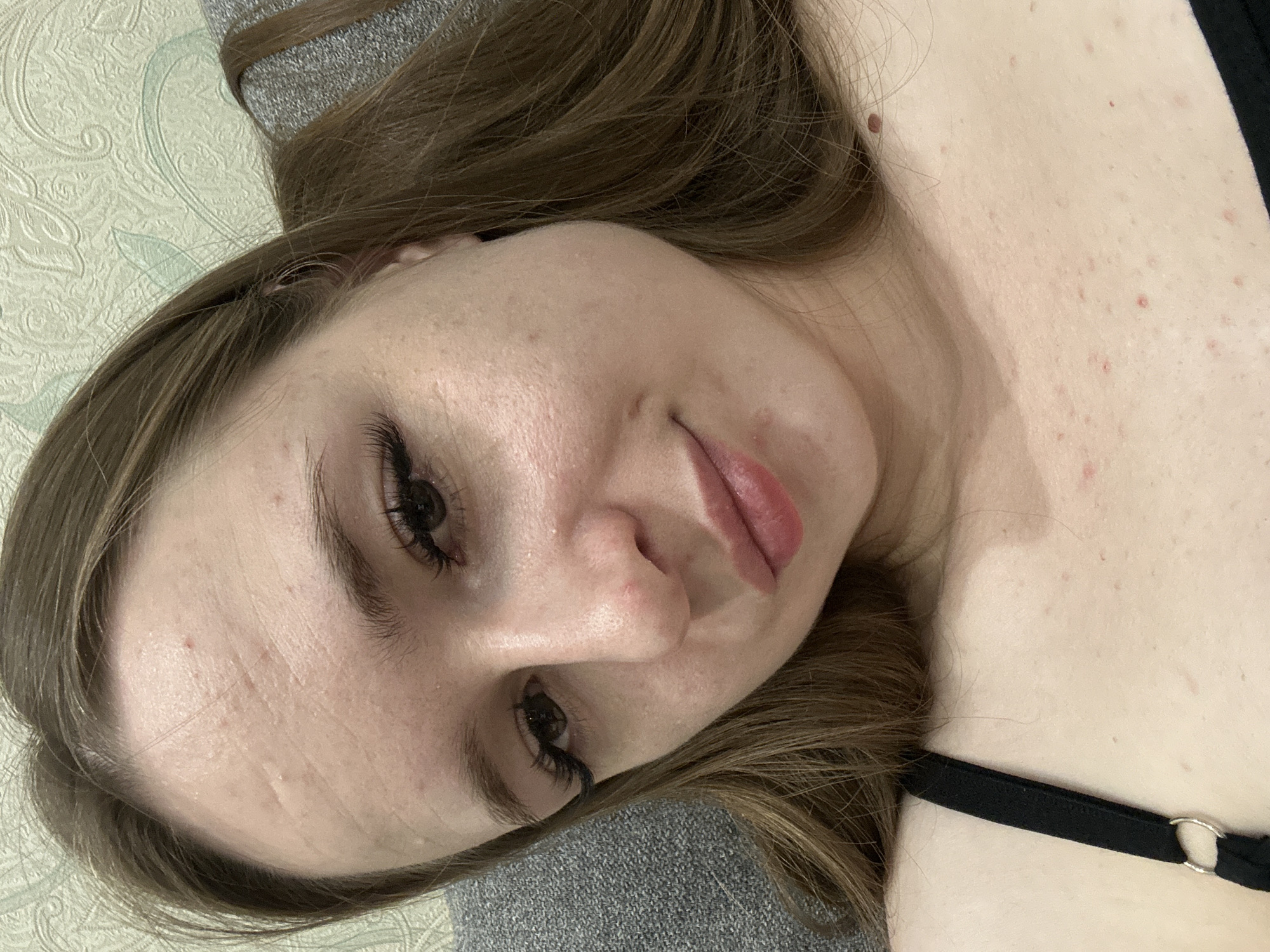 A close-up, indoor, selfie-style photo features a young woman with long, brown hair. She is wearing a black bra strap visible on her left side. Her face is partially visible, with her forehead, eyes, nose, and mouth in focus. The background appears to be an indoor setting with a textured wallpaper and a grey surface, possibly a sofa. The woman has fair skin with some blemishes and what appear to be extensions on her eyelashes. She is looking directly at the camera with a slight smile. The lighting is soft, and the overall image quality is clear. The location is unknown.