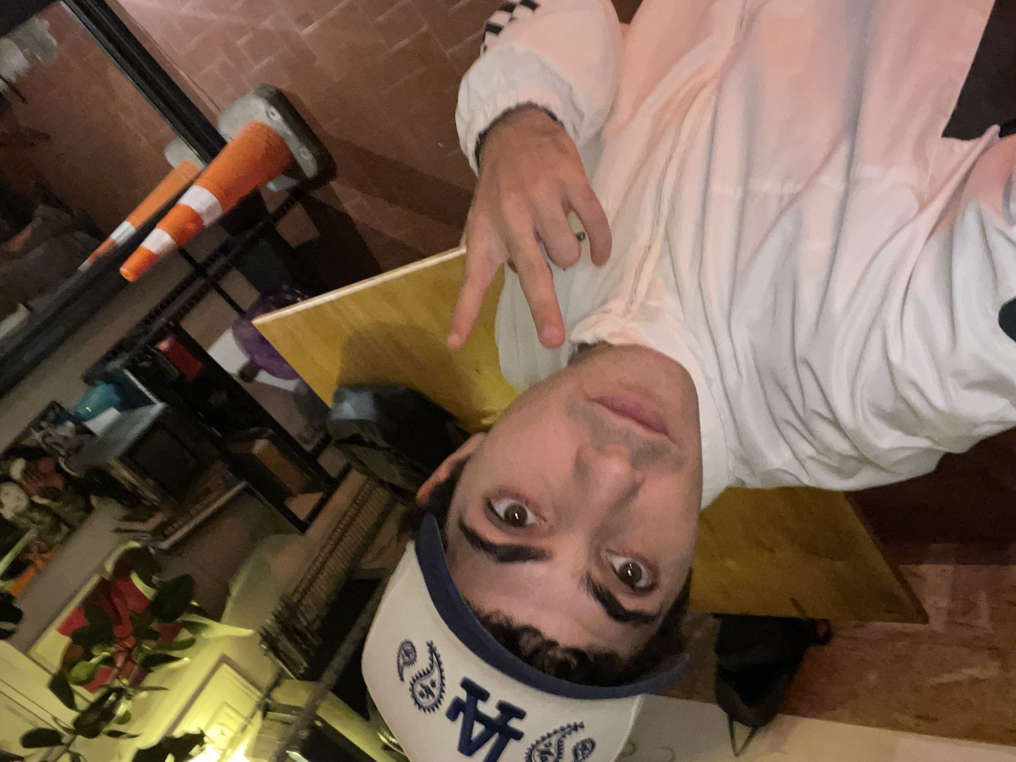 The image is a rotated selfie, capturing a young person (male-presenting) in a casual setting. The subject is centered in the frame, looking directly into the camera with a neutral expression. He has brown eyes and dark, curly hair visible under a white baseball cap with a blue brim. The cap is embroidered with blue paisley patterns and a prominent stylized "LA" logo.
He is wearing a white jacket or top, which features black and white checkered stripes on the sleeve of his left arm. His left hand is raised near his face, making a hand gesture with his index and middle fingers extended upwards, commonly known as a 'peace' or 'V' sign.
The background is somewhat blurred but offers contextual details. To the far left, two orange and white traffic cones are visible, suggesting an urban or industrial-themed environment. The setting appears to be indoors or a semi-covered space, possibly a café, bar, or shop, indicated by a reddish-brown brick wall, shelving units with various objects, and what looks like potted plants, one with reddish leaves. A wooden surface, likely a table or counter, stretches across the mid-ground. The lighting is artificial, suggesting the photo was taken in the evening or an indoor setting. The overall mood is informal.