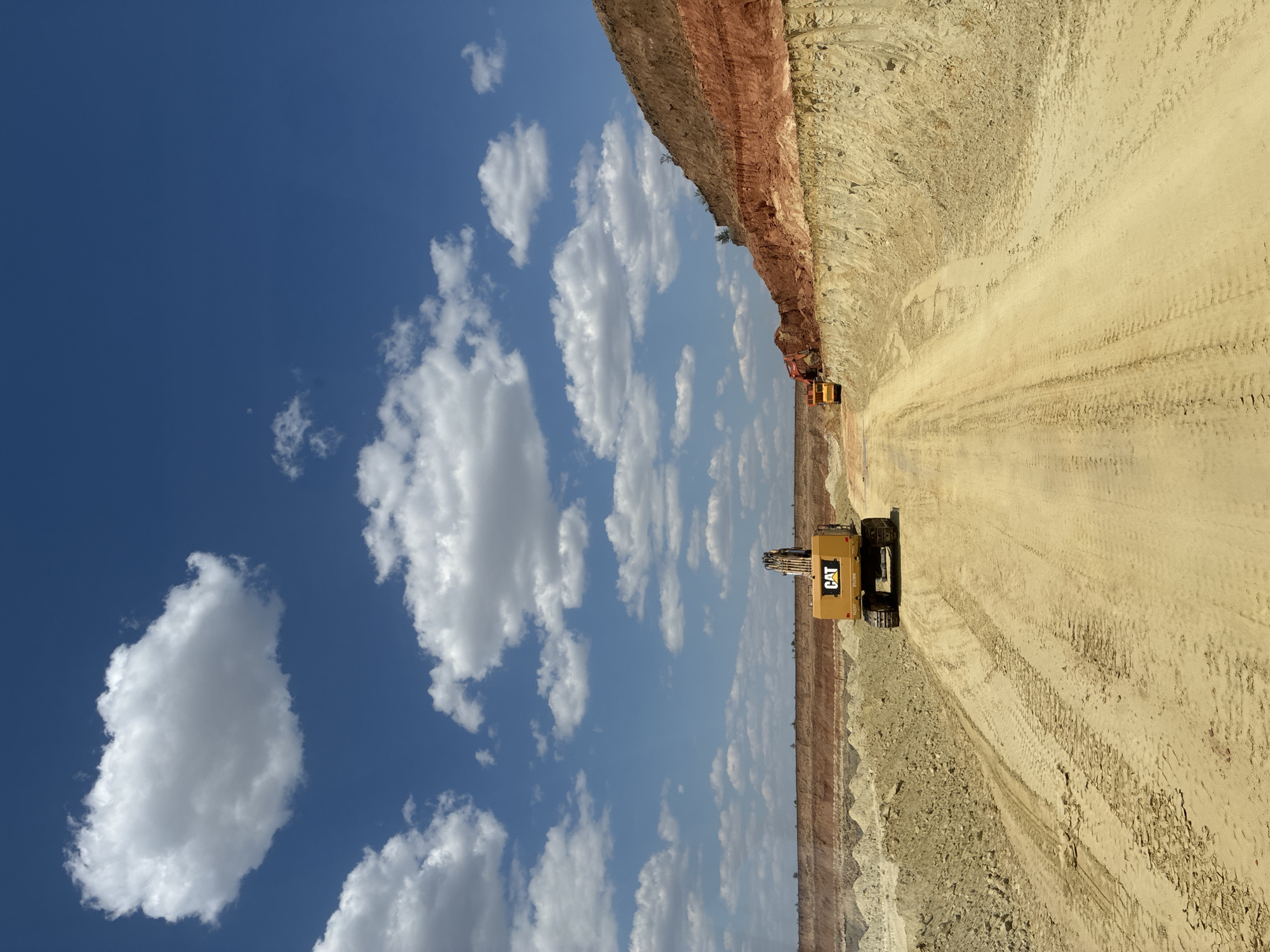 An outdoor, daytime, wide-angle shot captures a bright blue sky filled with fluffy white clouds over a large excavation site. The ground is sandy and appears to be a dirt road with tire tracks visible. On the left side of the road, a yellow CAT excavator is parked. Further down the road, another yellow construction vehicle, possibly a dump truck, is partially visible. The excavation site features layered soil in shades of tan, yellow, and reddish-brown, indicating different strata of earth. The overall scene depicts an active or recently active construction or mining operation under a clear sky.
