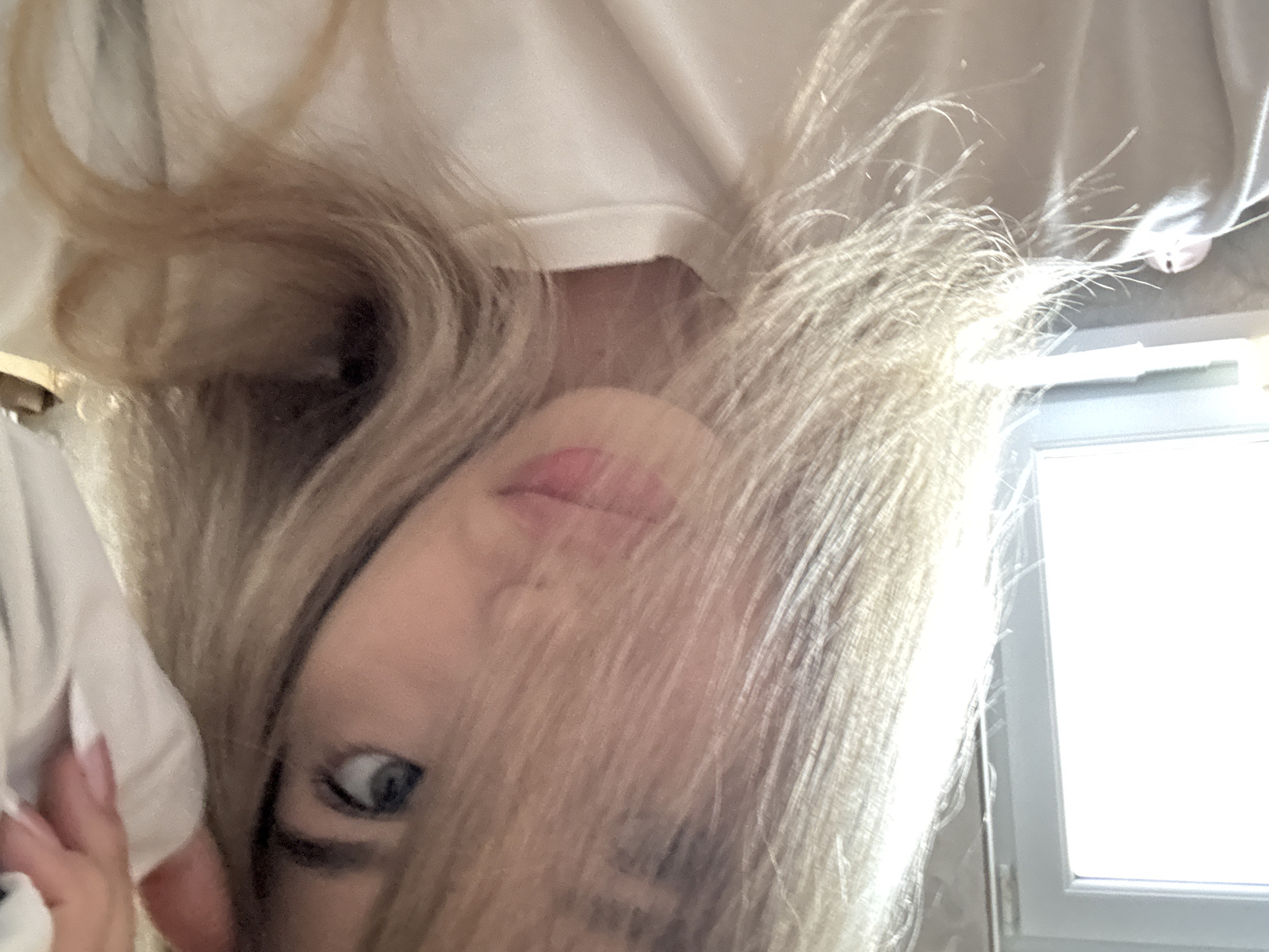 A close-up, upside-down perspective captures a woman with blonde hair partially obscuring her face. Her lips are visible, painted a light pink. One blue eye is visible, looking directly at the camera. She wears a white garment, likely a shirt or blouse. In the background, a white window frame and a window filled with bright light are visible. The location is not discernible based solely on the image.