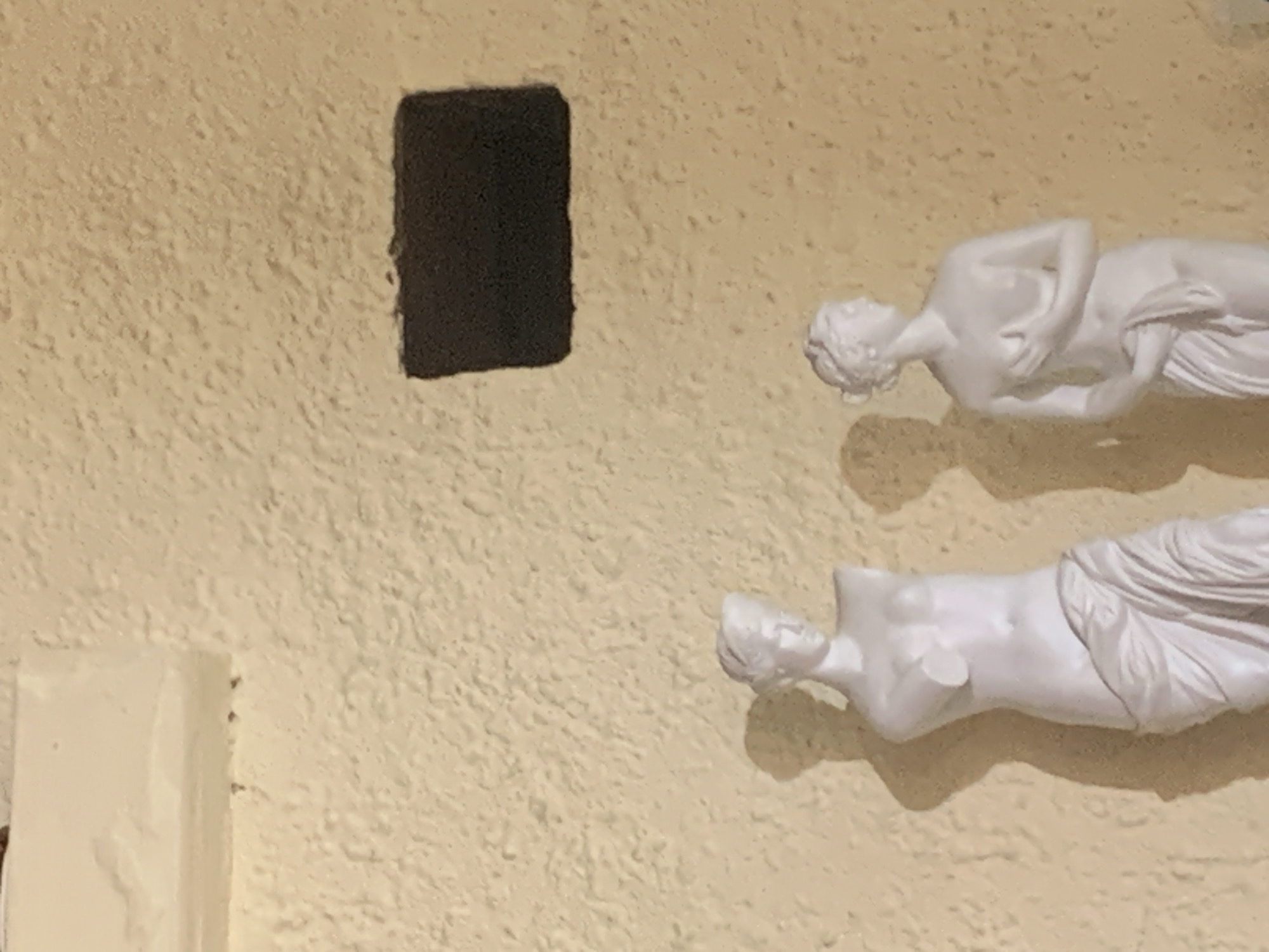 The image depicts two plaster statues of women against a textured, pale yellow wall. The statue on the left is headless and partially draped in fabric. The statue on the right is complete, with its head turned to the side, and its hands covering its chest. A dark, rectangular opening is visible in the upper right quadrant of the wall. The lighting suggests an indoor setting with shadows cast by the statues. The location in the city of Sergeevo, Russia, is not visually evident in the image.
