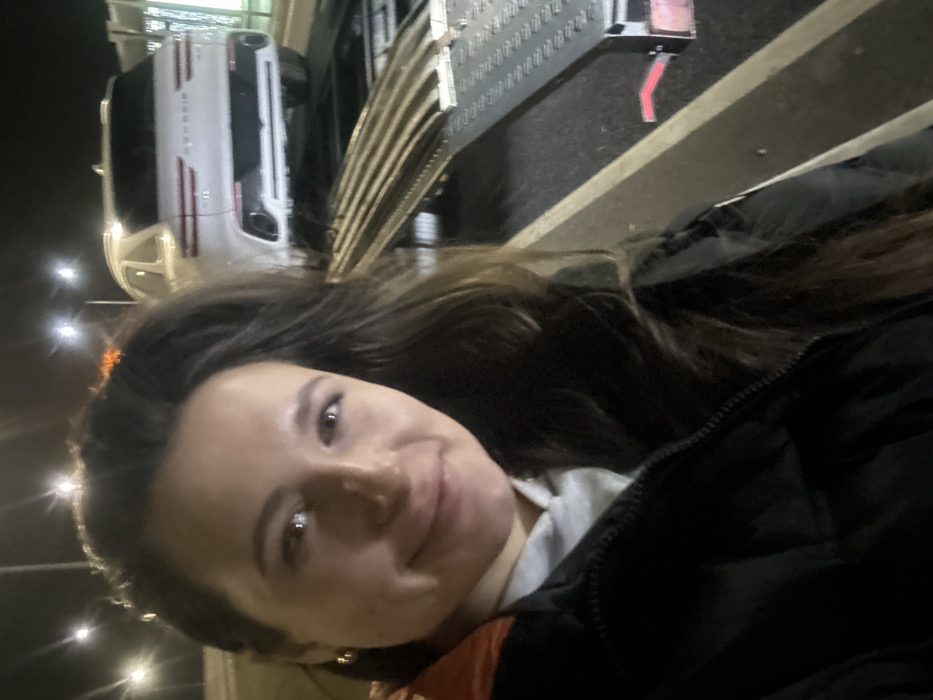 A woman with long brown hair smiles at the camera. She wears a black jacket over a light-colored shirt. In the background, a white SUV is parked on a ramp next to a metal platform. The scene is lit by streetlights, indicating it's nighttime.
