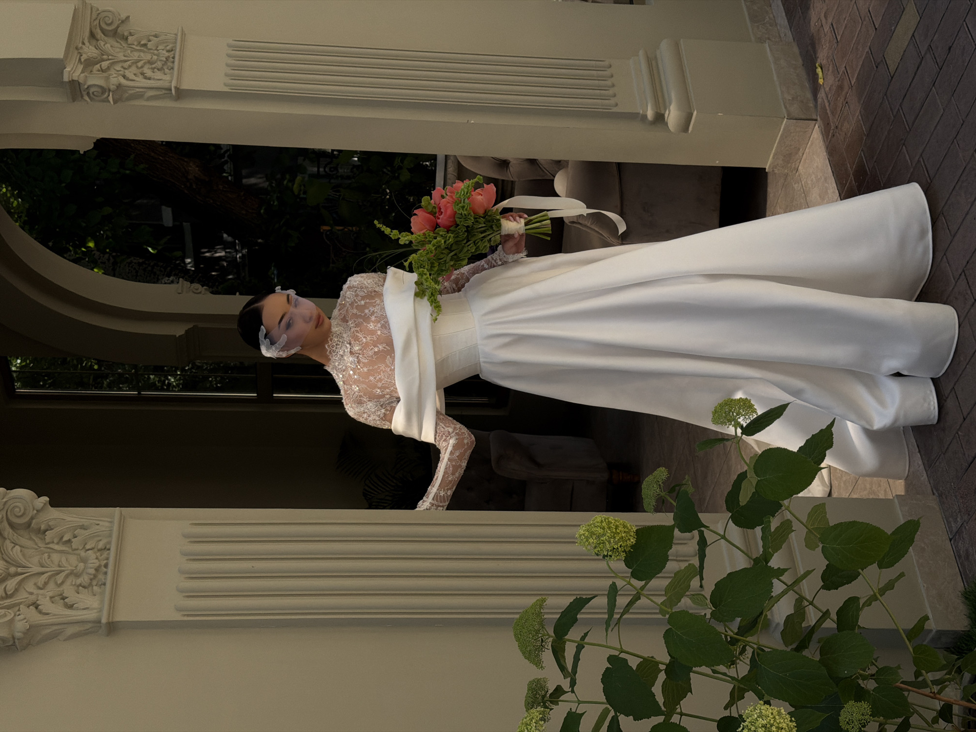 A woman in a wedding dress stands in what appears to be a doorway or entrance in Bishkek City, Kyrgyzstan. She is holding a bouquet of flowers. The doorway is part of a building with ornate architectural details. The background includes greenery. The flooring outside the doorway is brick.